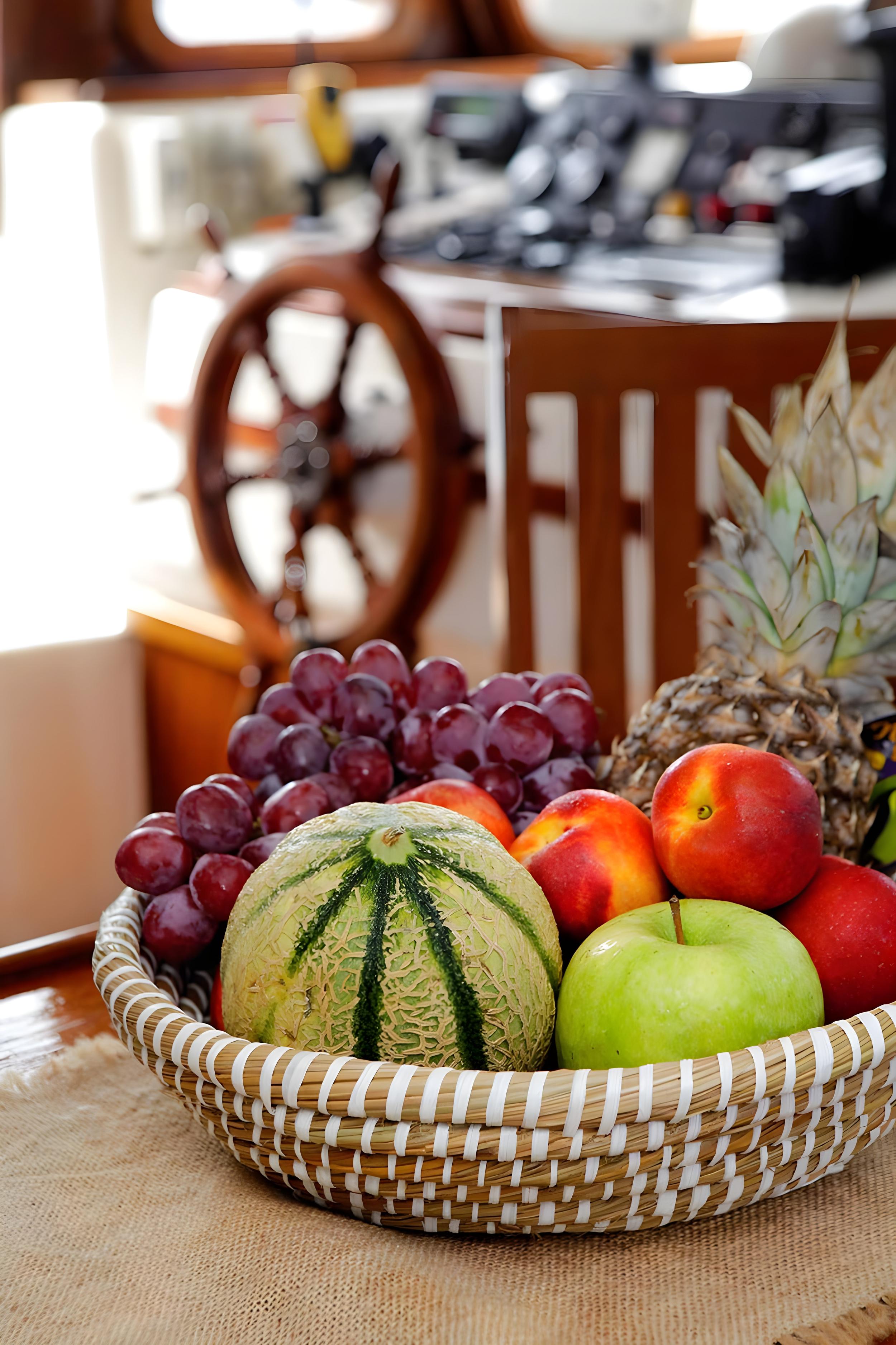 Woven basket of fresh fruit including melon, grapes, apples and pineapple on gulet with wooden helm wheel visible behind