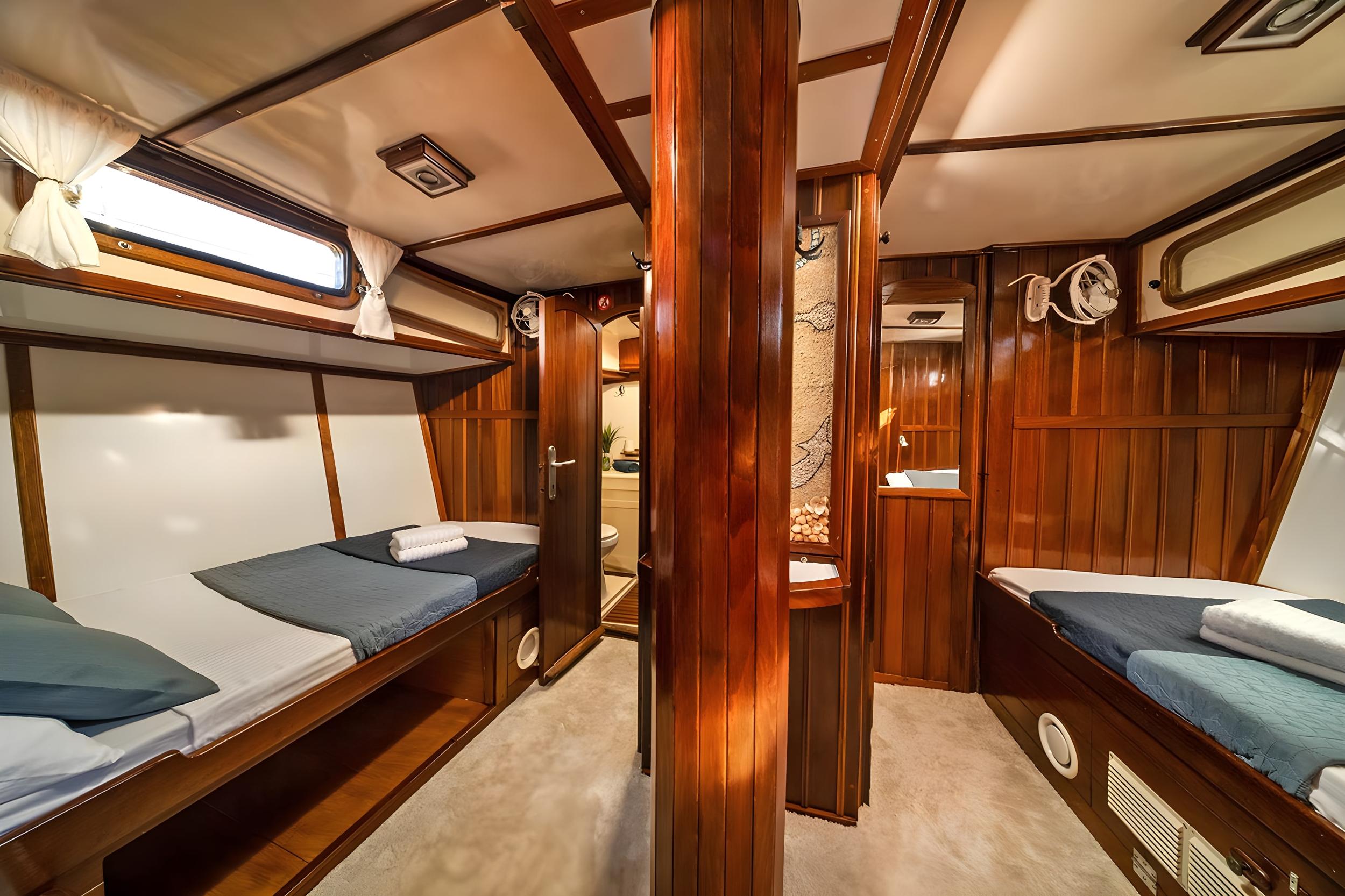 Gulet twin cabin with two single beds, upper bunk, warm mahogany wood panelling, and en-suite shower room visible through open door