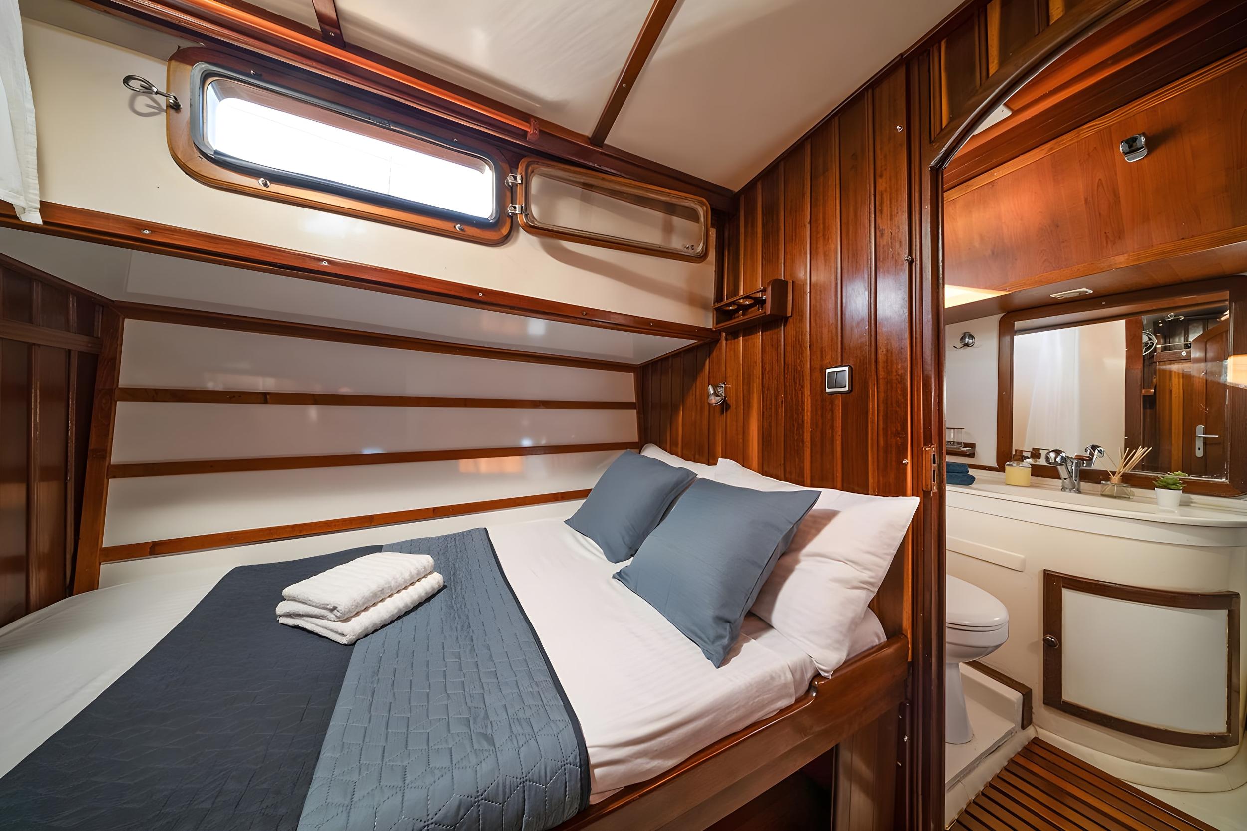 Gulet double cabin with grey quilted bedspread, two blue pillows, folded towels, warm teak panelling, and open en-suite showing basin and toilet