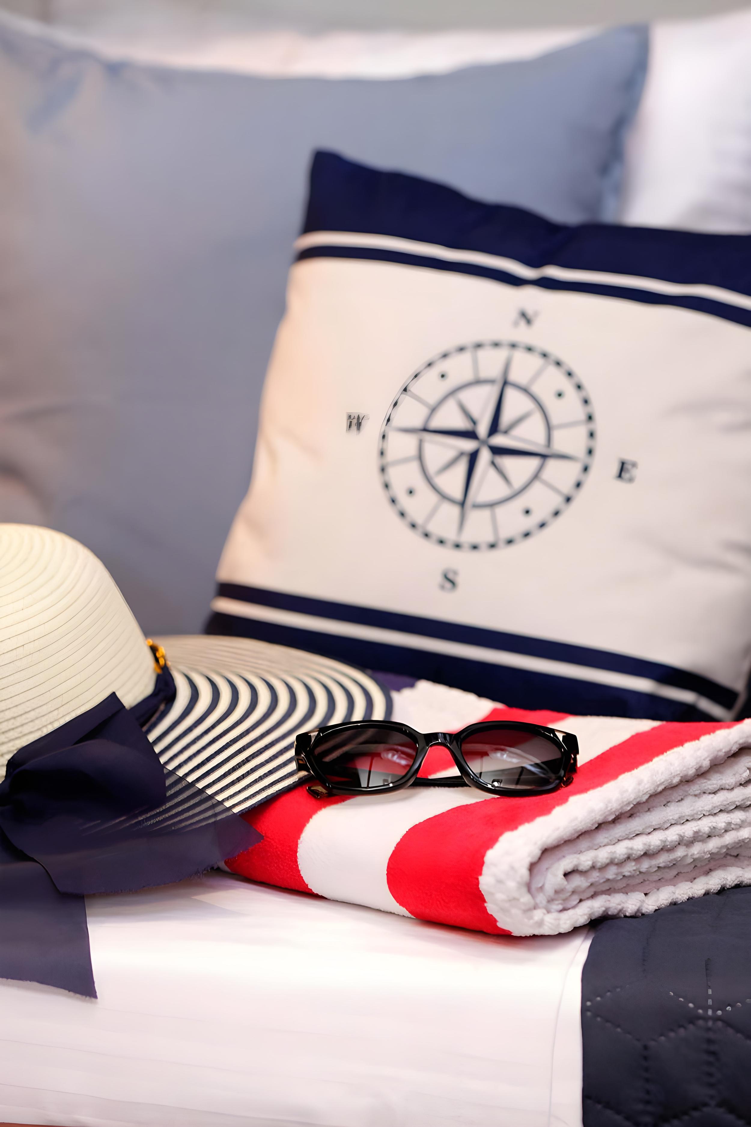Gulet cabin bed dressed with navy compass rose cushion, red-striped beach towel, striped sun hat, and black sunglasses