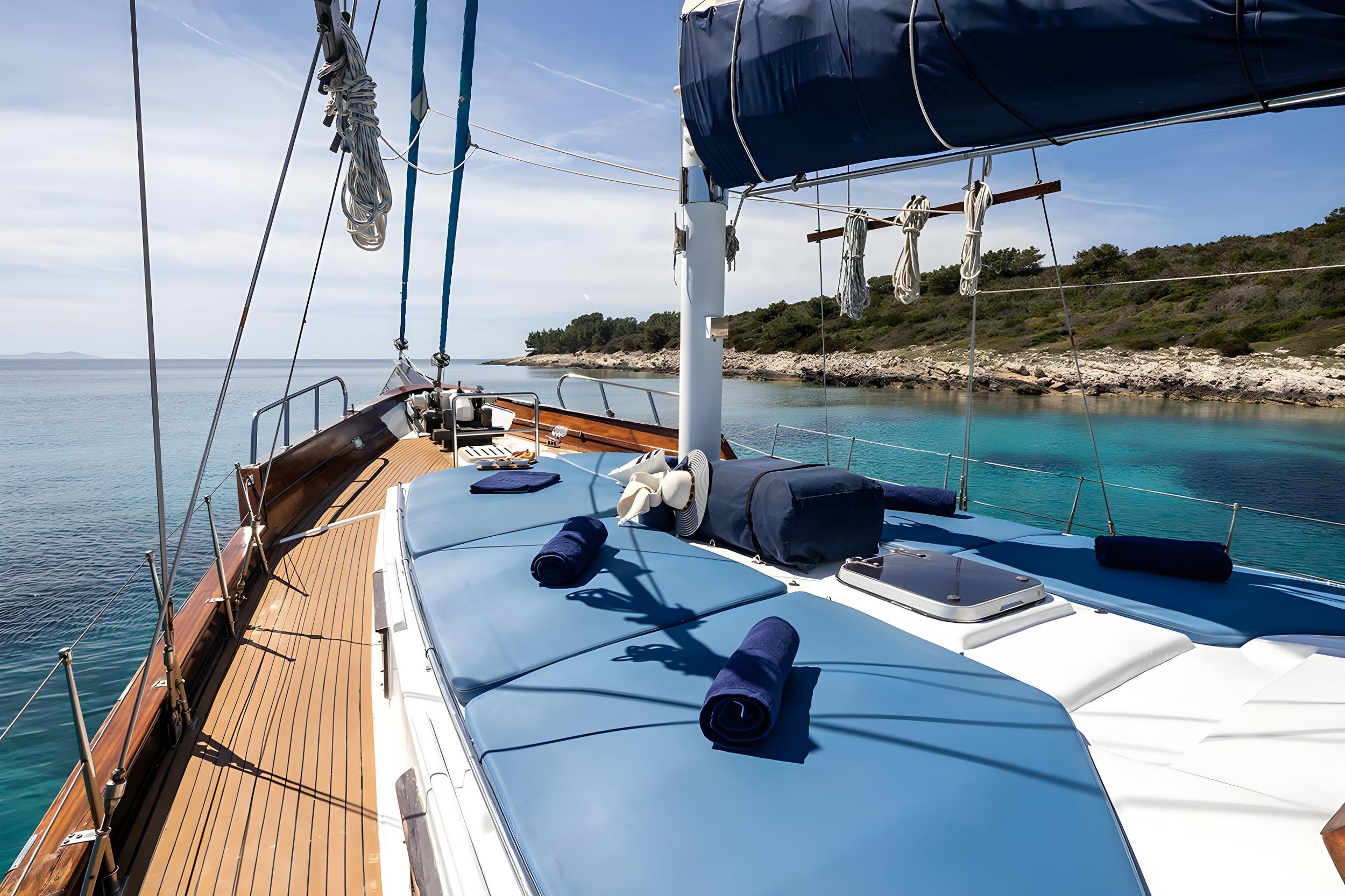 Gulet bow sun deck with blue cushioned sunbathing pads, rolled navy towels, teak side decks, and turquoise Croatian coastline anchorage