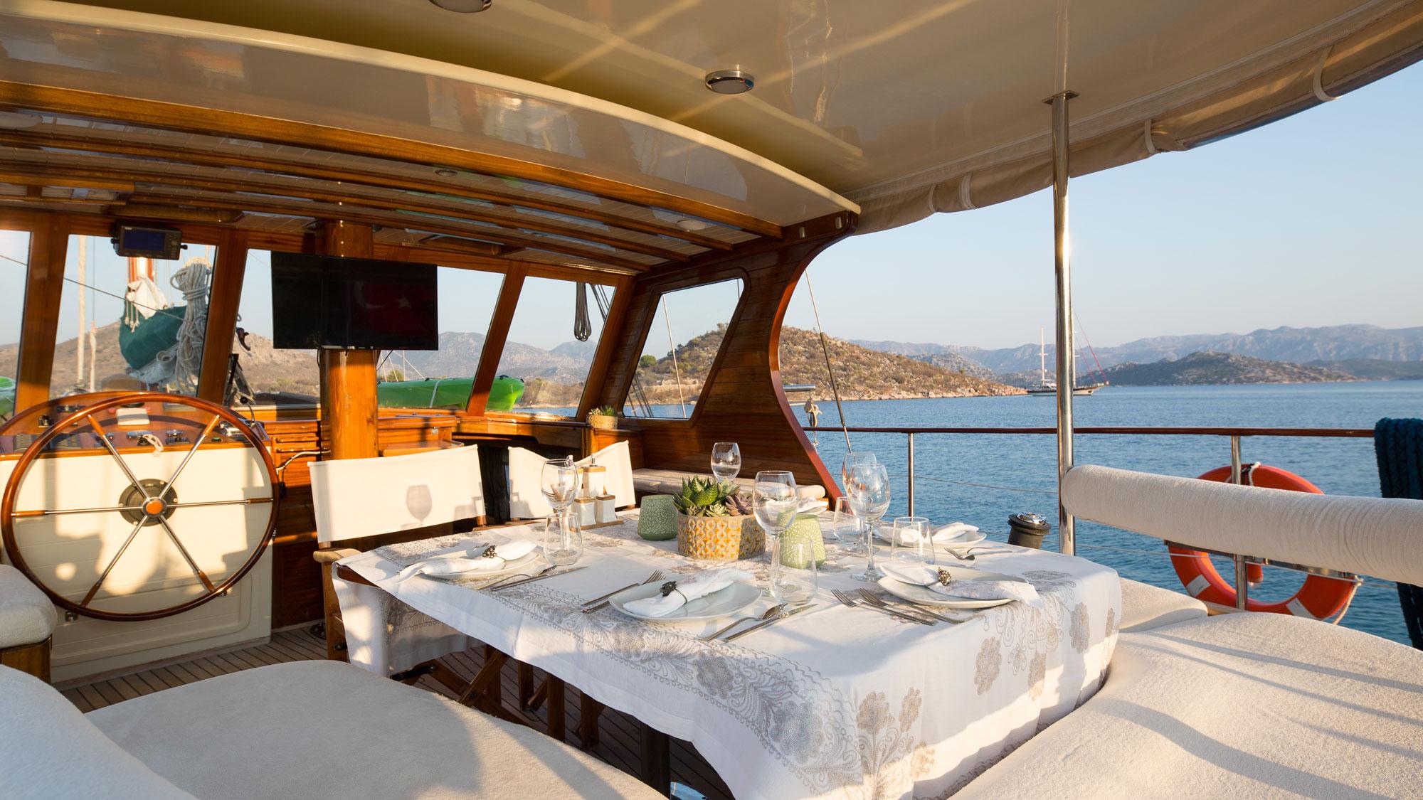 covered aft deck with formal dining table, ship's wheel, and panoramic windows overlooking coastal waters