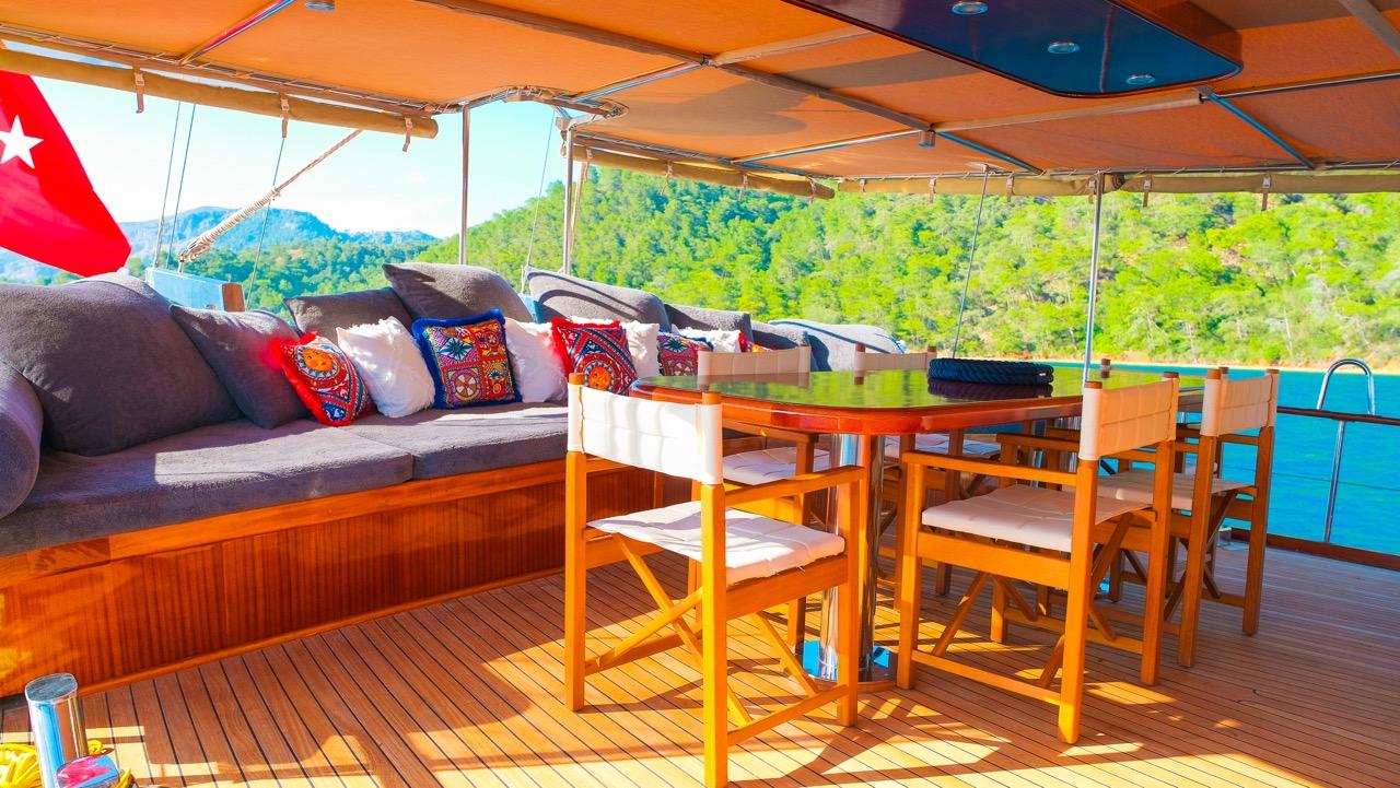 Gulet aft deck with built-in seating, wooden dining table, chairs and decorative cushions
