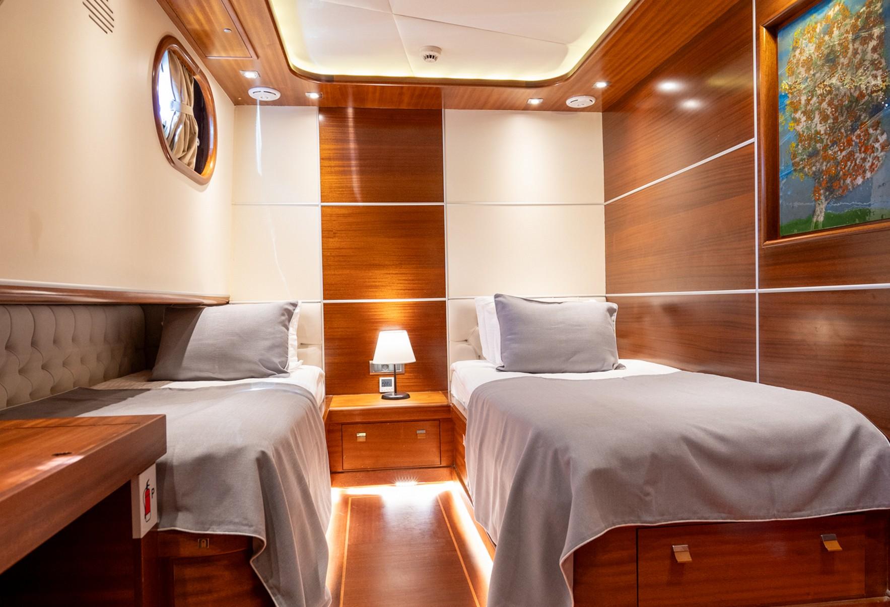 twin guest cabin with two single beds, wooden paneling, bedside table with lamp, and porthole window