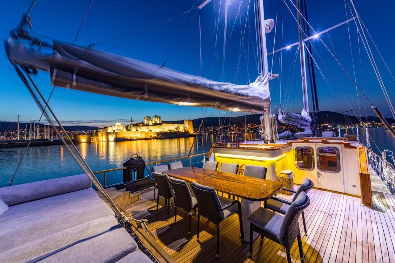 gulet aft deck with teak dining table and chairs at twilight, illuminated castle fortress in background