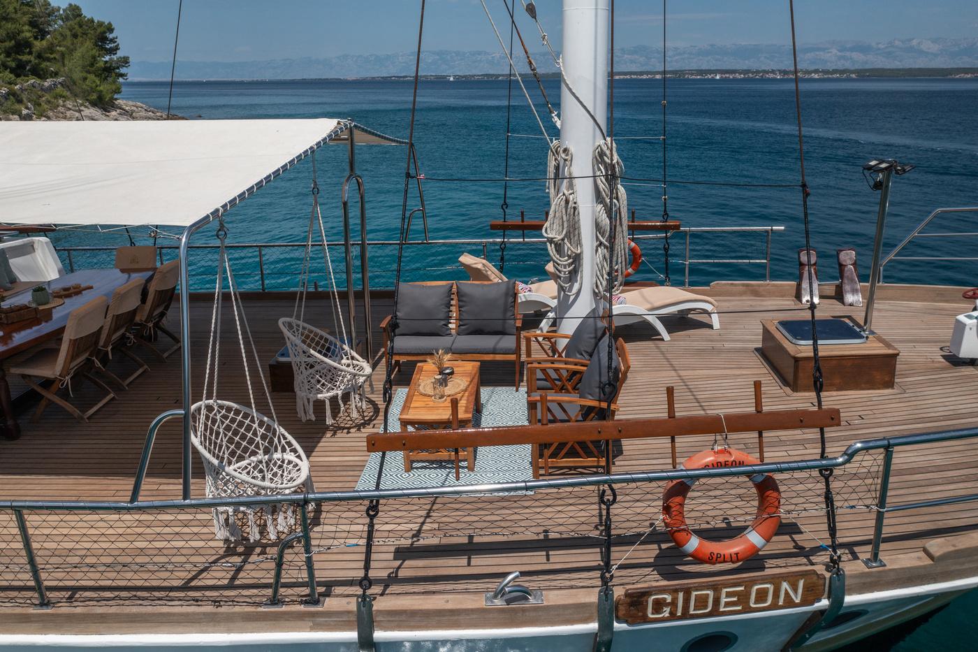 gulet deck featuring teak flooring, built-in seating with cushions, central mast with rigging, and hanging swing chairs
