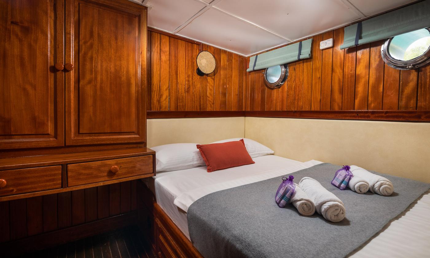 yacht guest cabin featuring double bed with white linens, rich wood paneling, portholes and built-in wardrobes