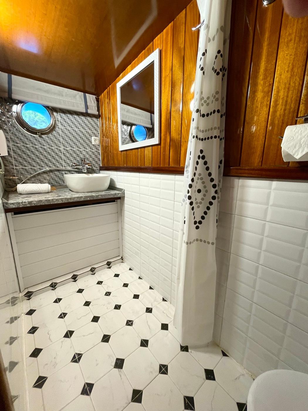 yacht bathroom with white vessel basin, marble countertop, teak cabinetry and two portholes