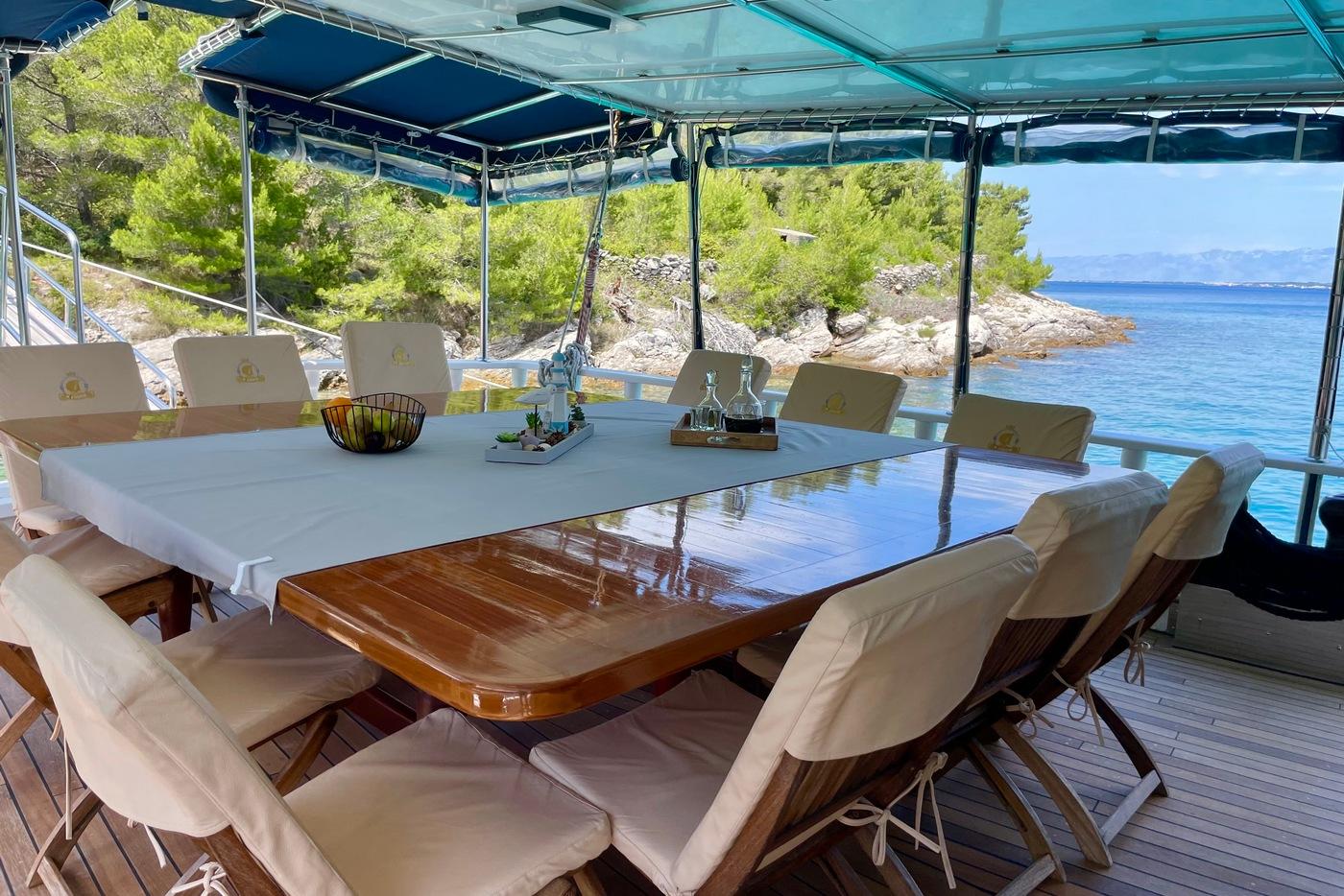 gulet aft deck dining table with cream chairs under awning overlooking Mediterranean coastline