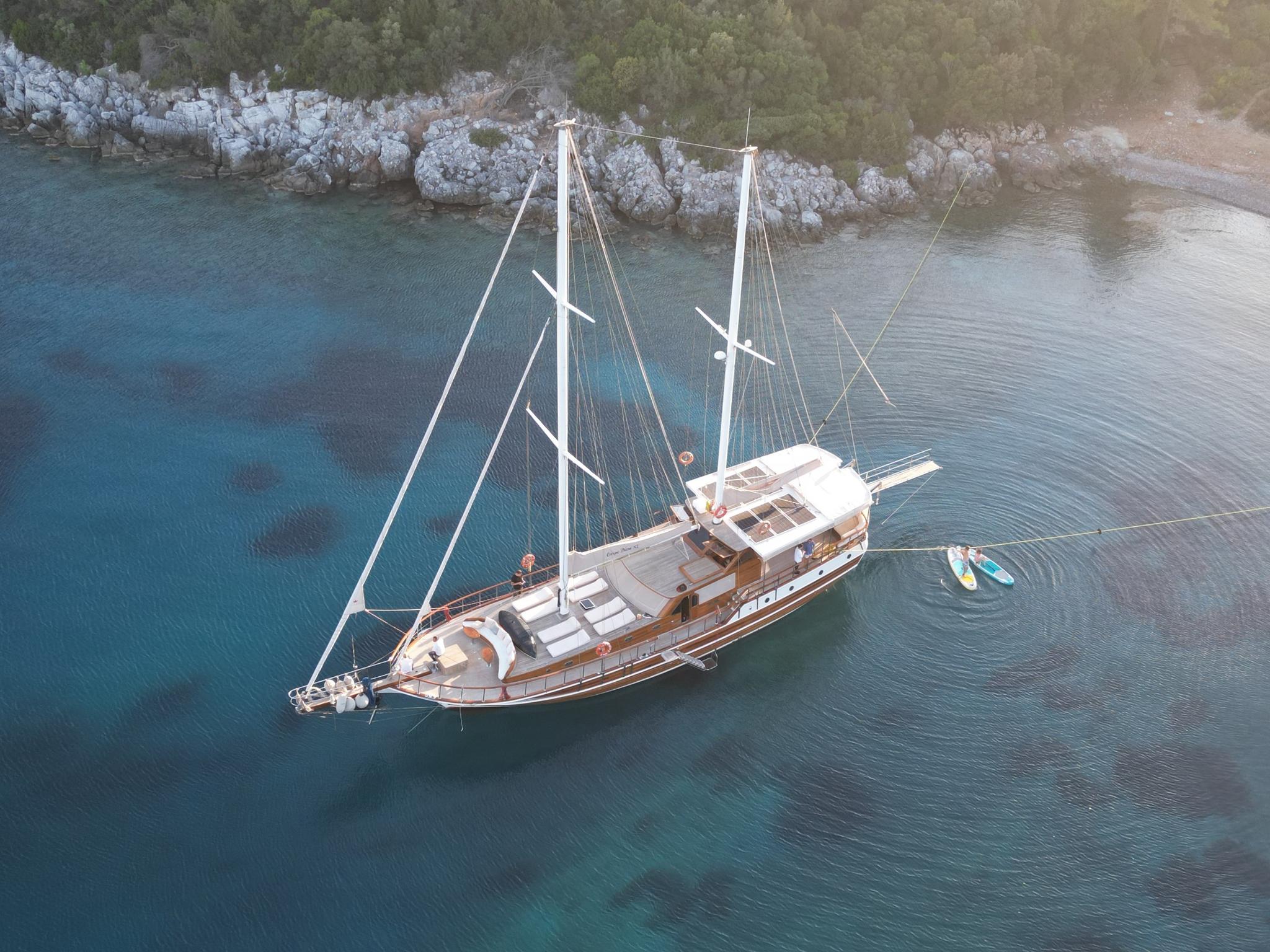 aerial view of white wooden gulet with two masts anchored in crystal clear turquoise waters near rocky coastline
