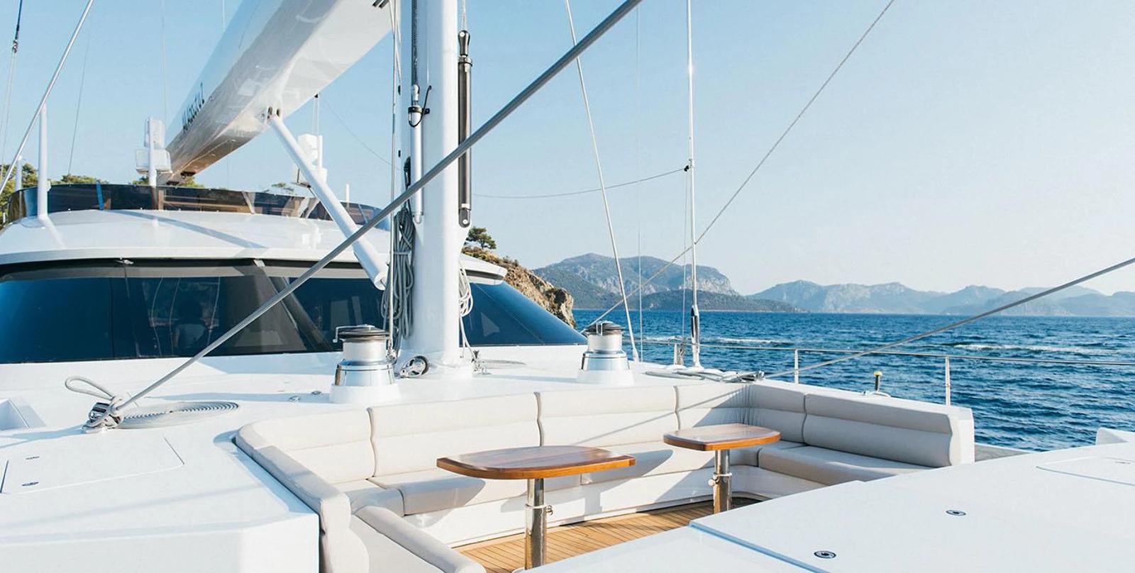 Bow deck seating area with white upholstered benches, teak table, and mountain coastline view