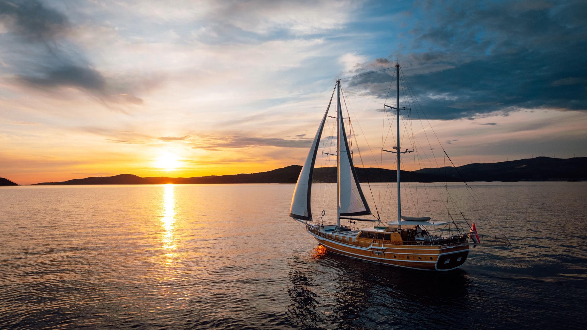 Traditional wooden gulet with white sails cruising at golden sunset with mountainous coastline