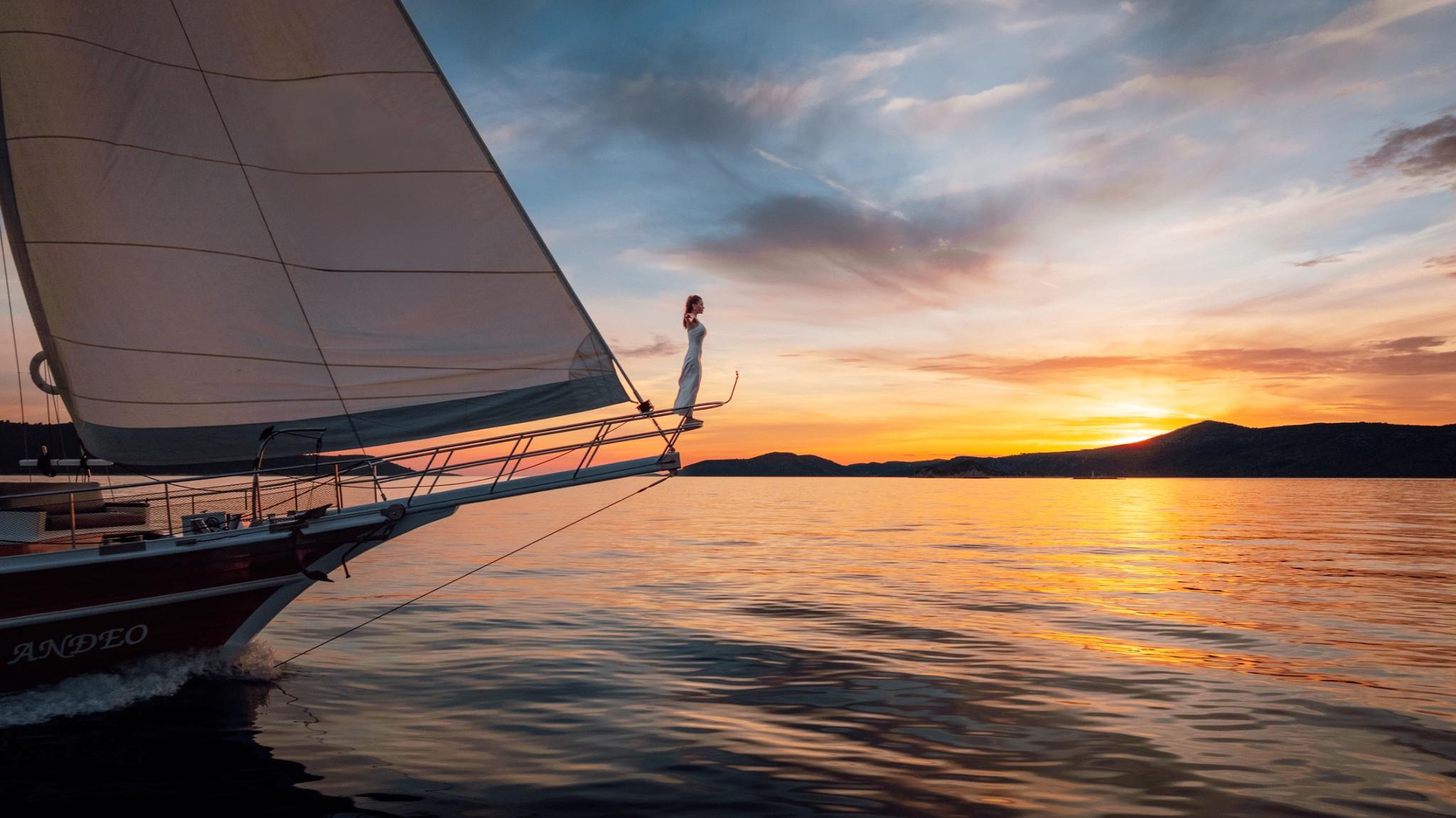 Traditional wooden gulet under sail at sunset with passenger standing on bowsprit
