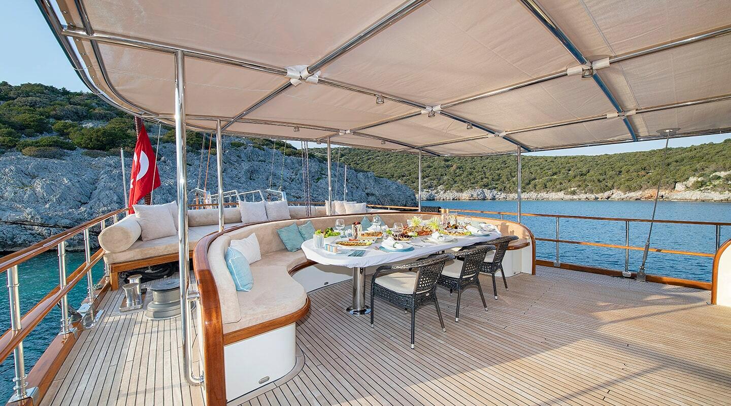 Yacht aft deck with curved seating, dining table and retractable awning overlooking coastline