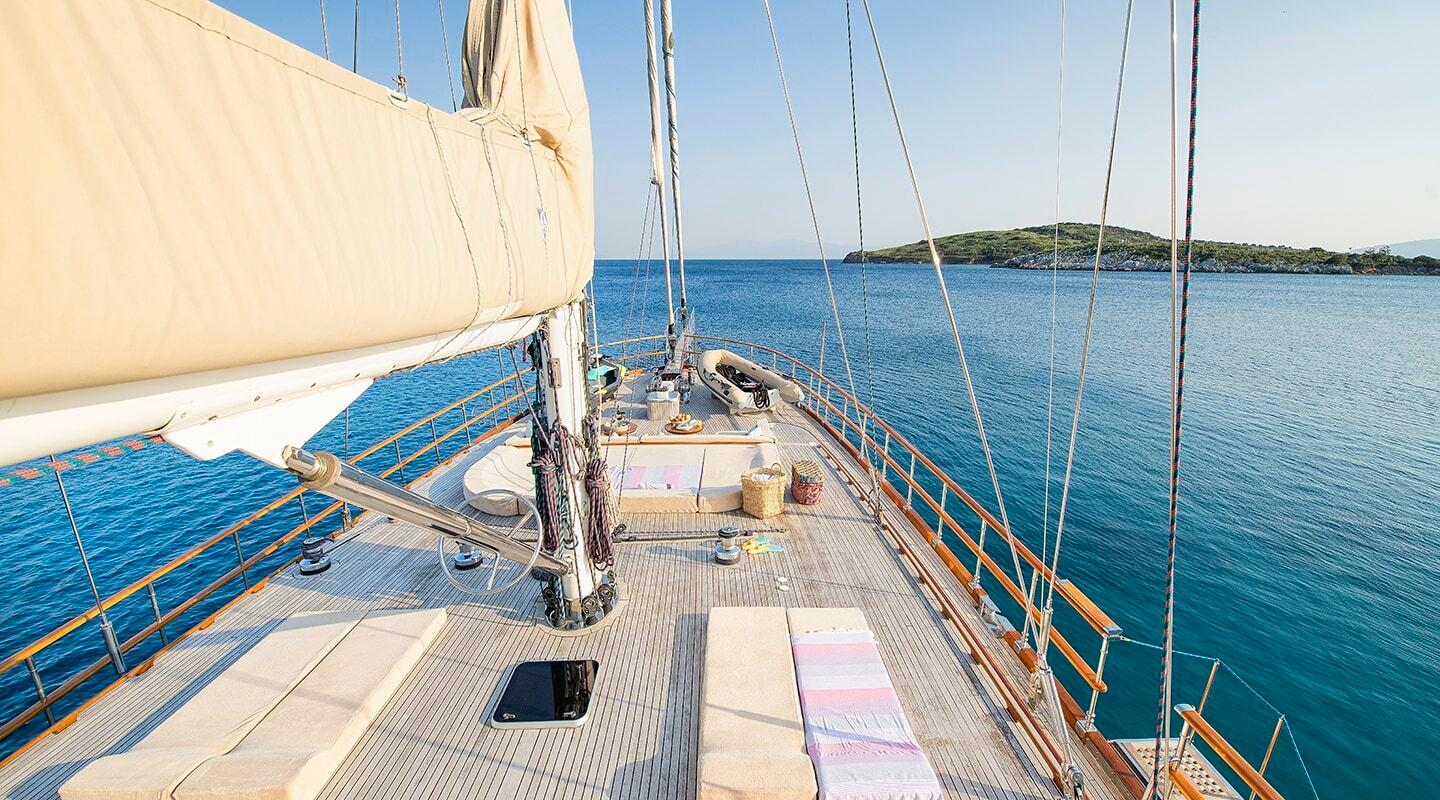 gulet bow deck with wooden planking, central mast, rigging and cream sails anchored in blue bay