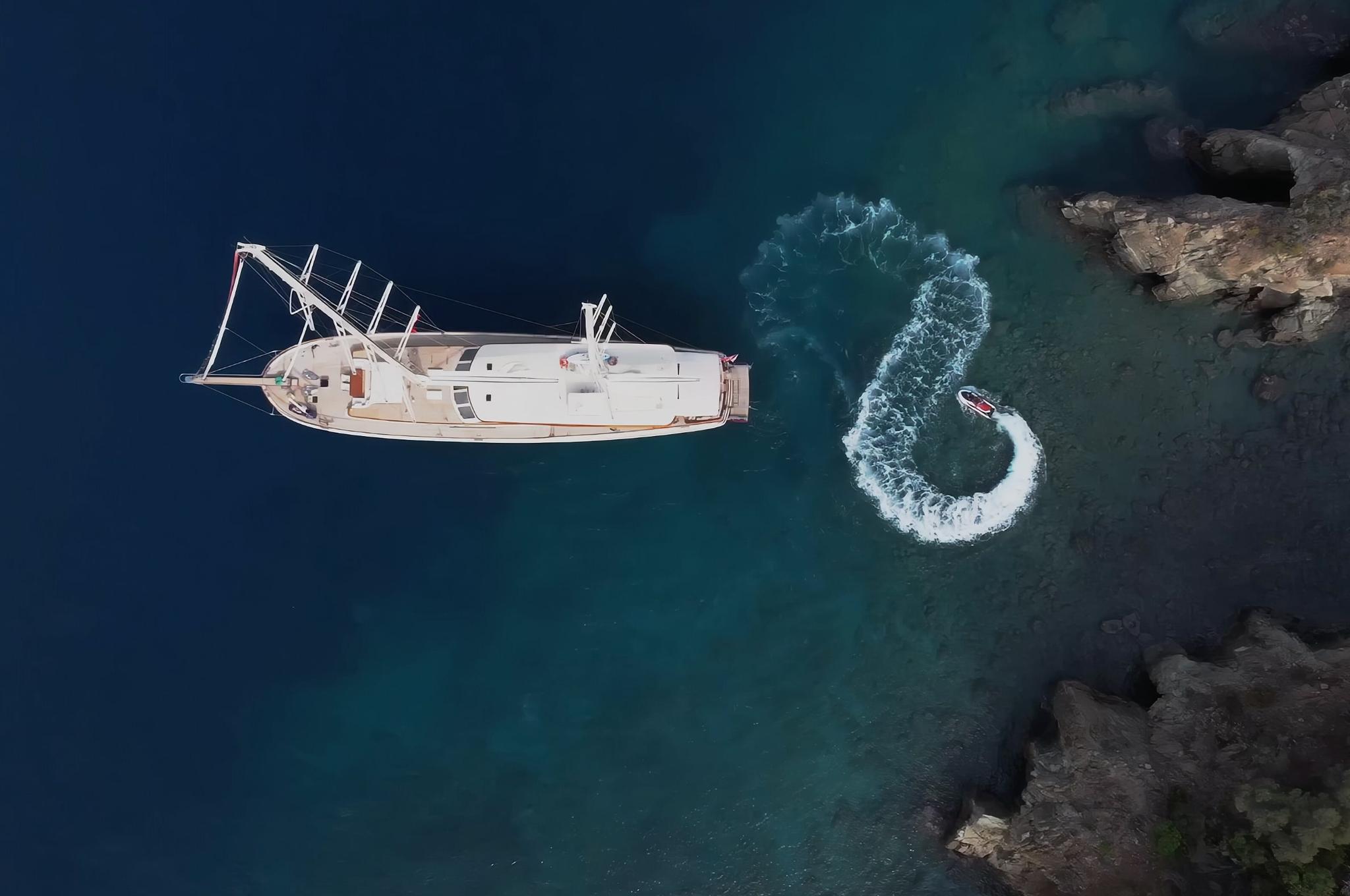 aerial view of white yacht anchored in turquoise waters near rocky coastline with jet ski creating wake