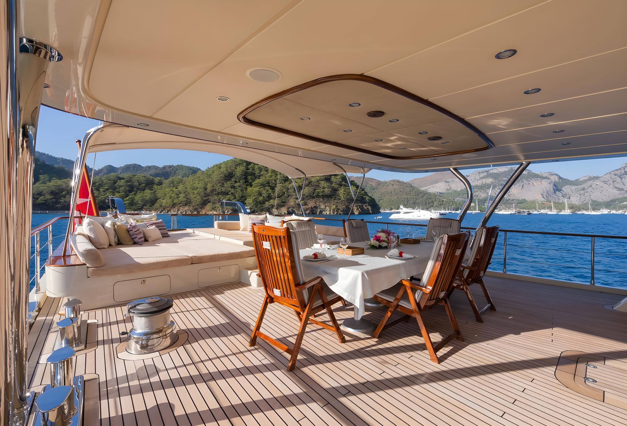 yacht aft deck with white dining table, teak chairs, cream lounger and mountain coastline views
