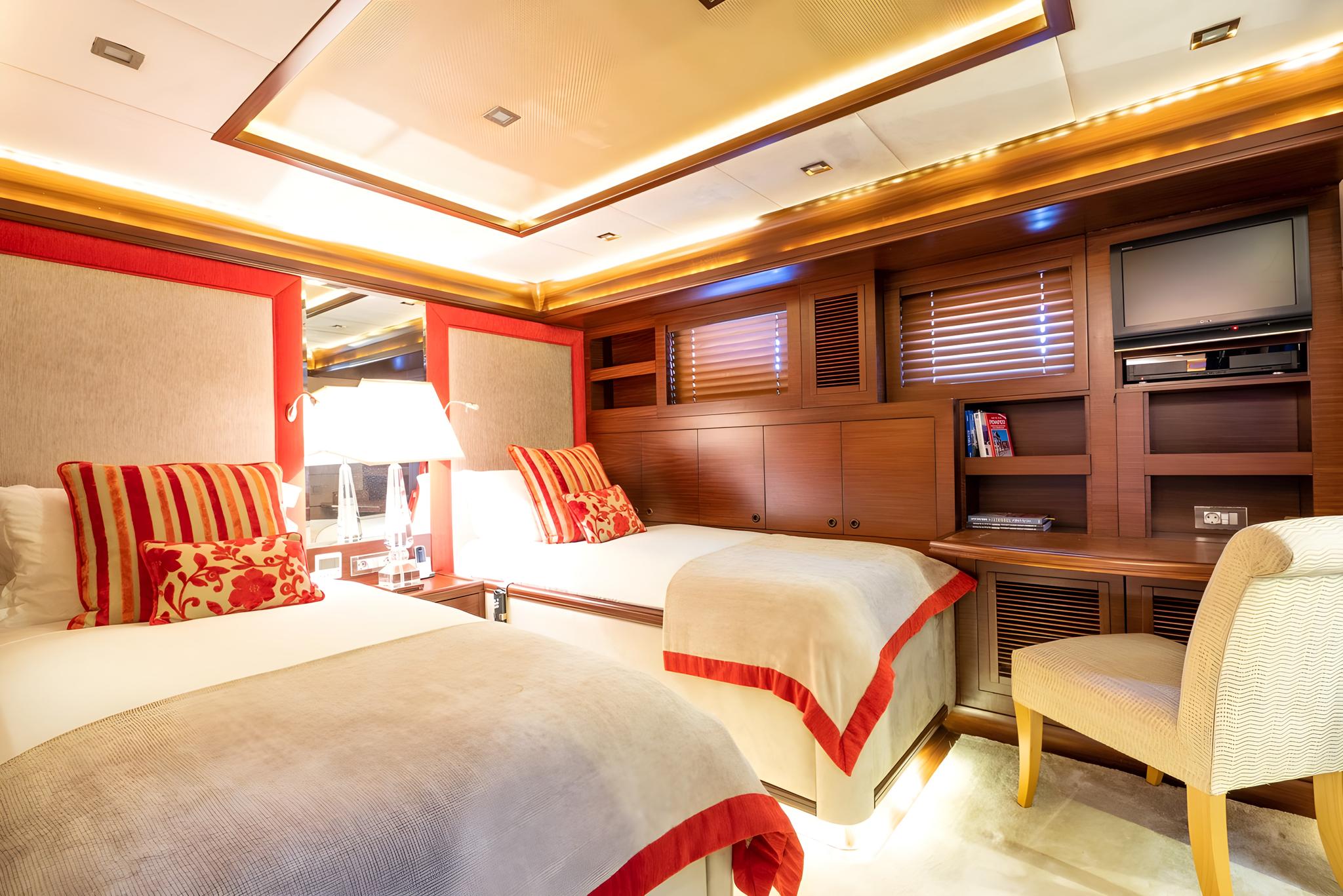 yacht guest cabin with twin beds, red striped pillows, built-in TV unit and wooden cabinetry