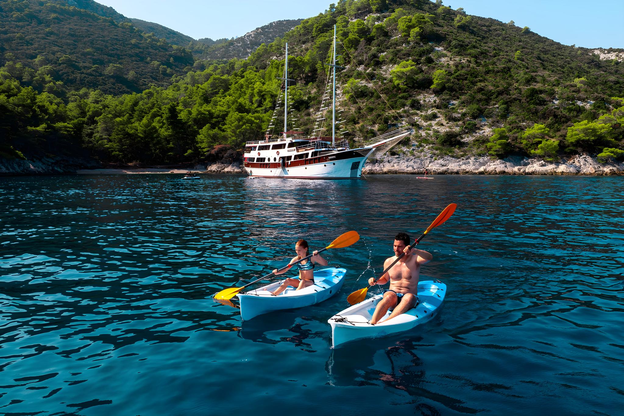 Two guests kayaking with orange paddles near luxury gulet in turquoise Mediterranean bay