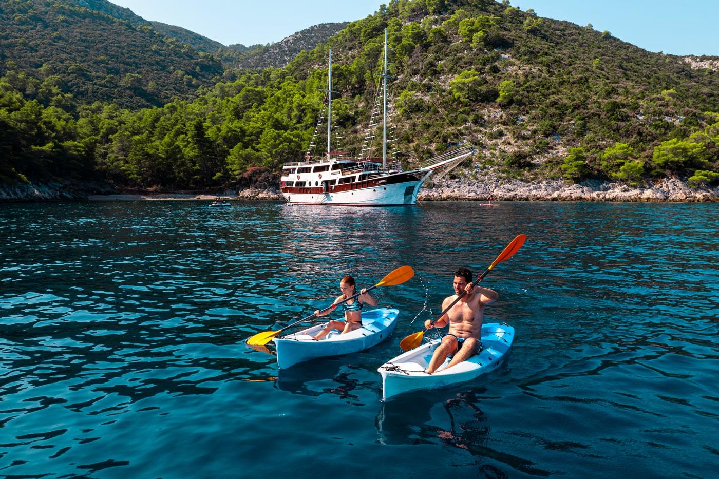 Two guests kayaking with orange paddles near luxury gulet in turquoise Mediterranean bay