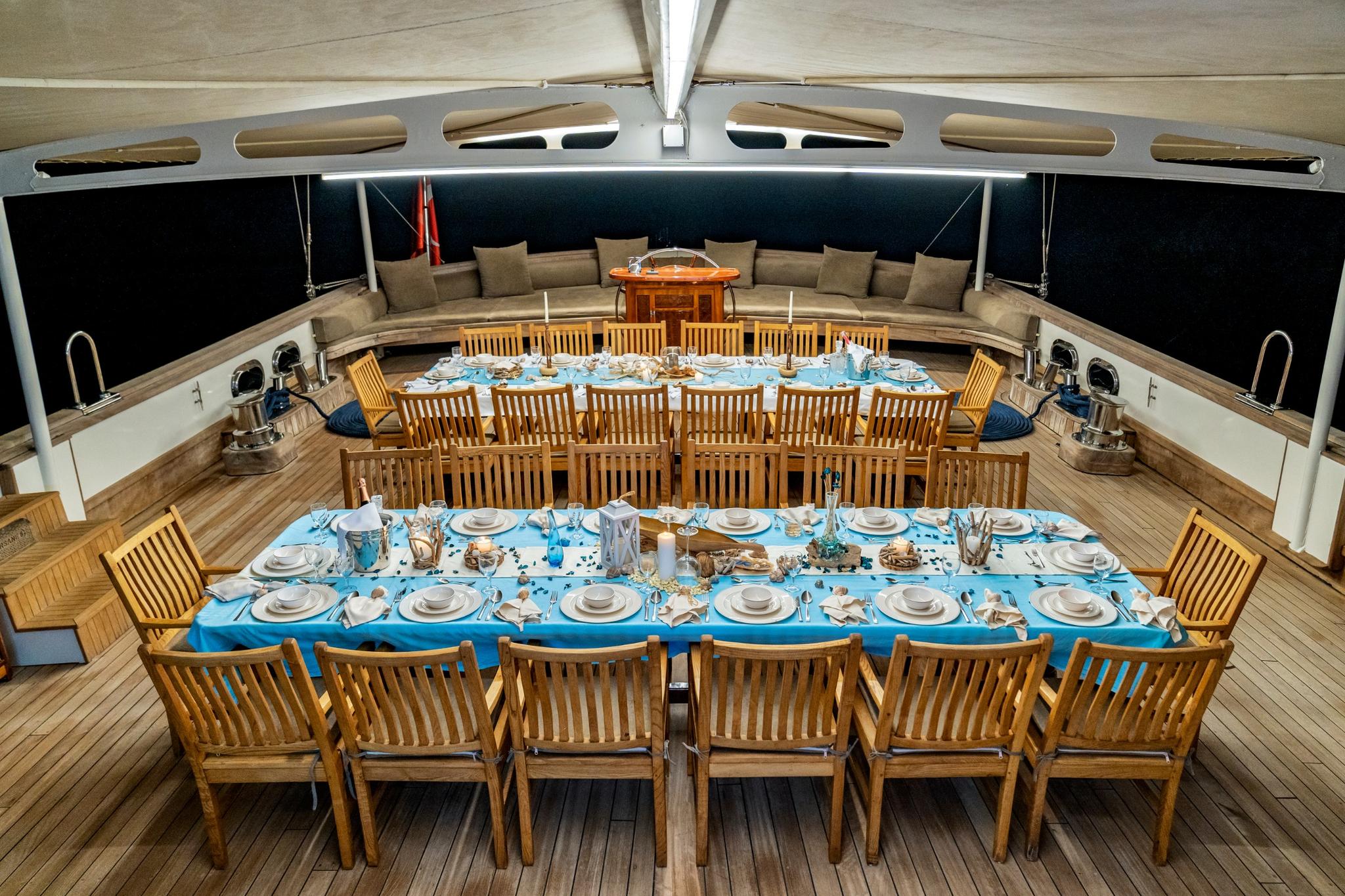 yacht aft deck with two formal dining tables set with blue tablecloths and teak chairs