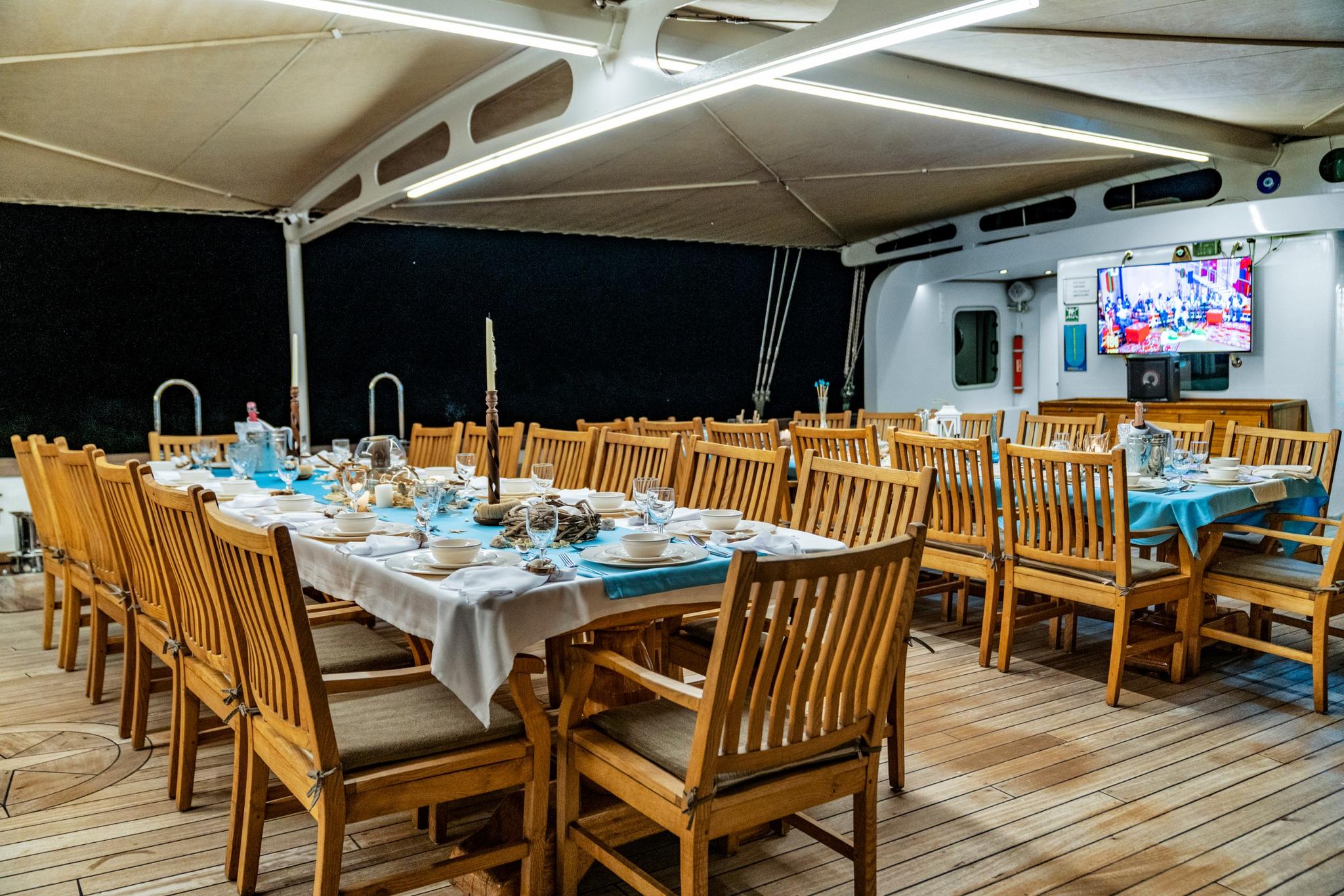 covered aft deck dining area with wooden chairs, white tablecloth and wall-mounted TV screen
