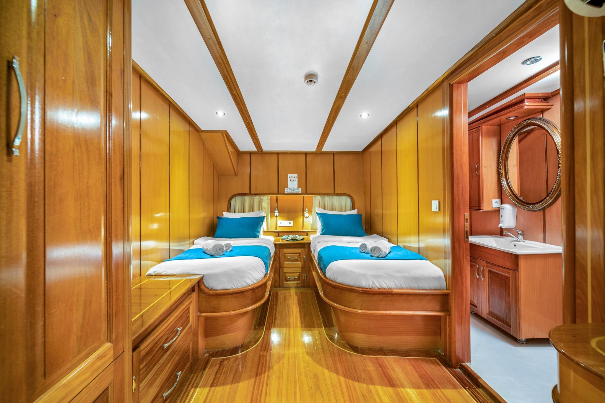 yacht twin cabin with polished wood paneling, two single beds with blue runners and en-suite bathroom