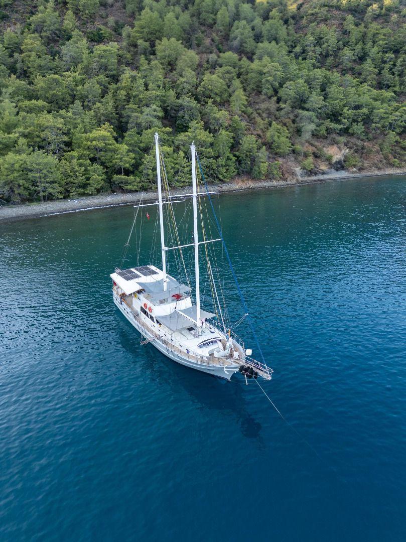 aerial view of white sailing yacht with two masts anchored in turquoise Mediterranean bay near forested coastline