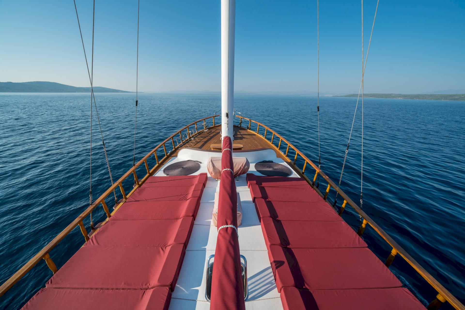 gulet bow deck with red cushioned loungers arranged around white mast on blue sea