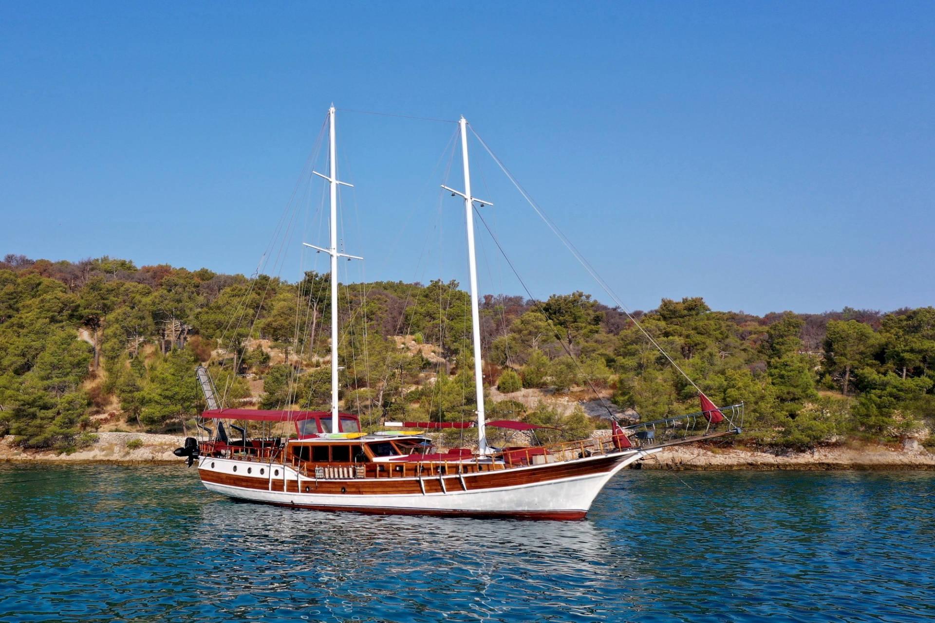 Traditional wooden gulet with two masts anchored in turquoise Mediterranean waters near forested coastline