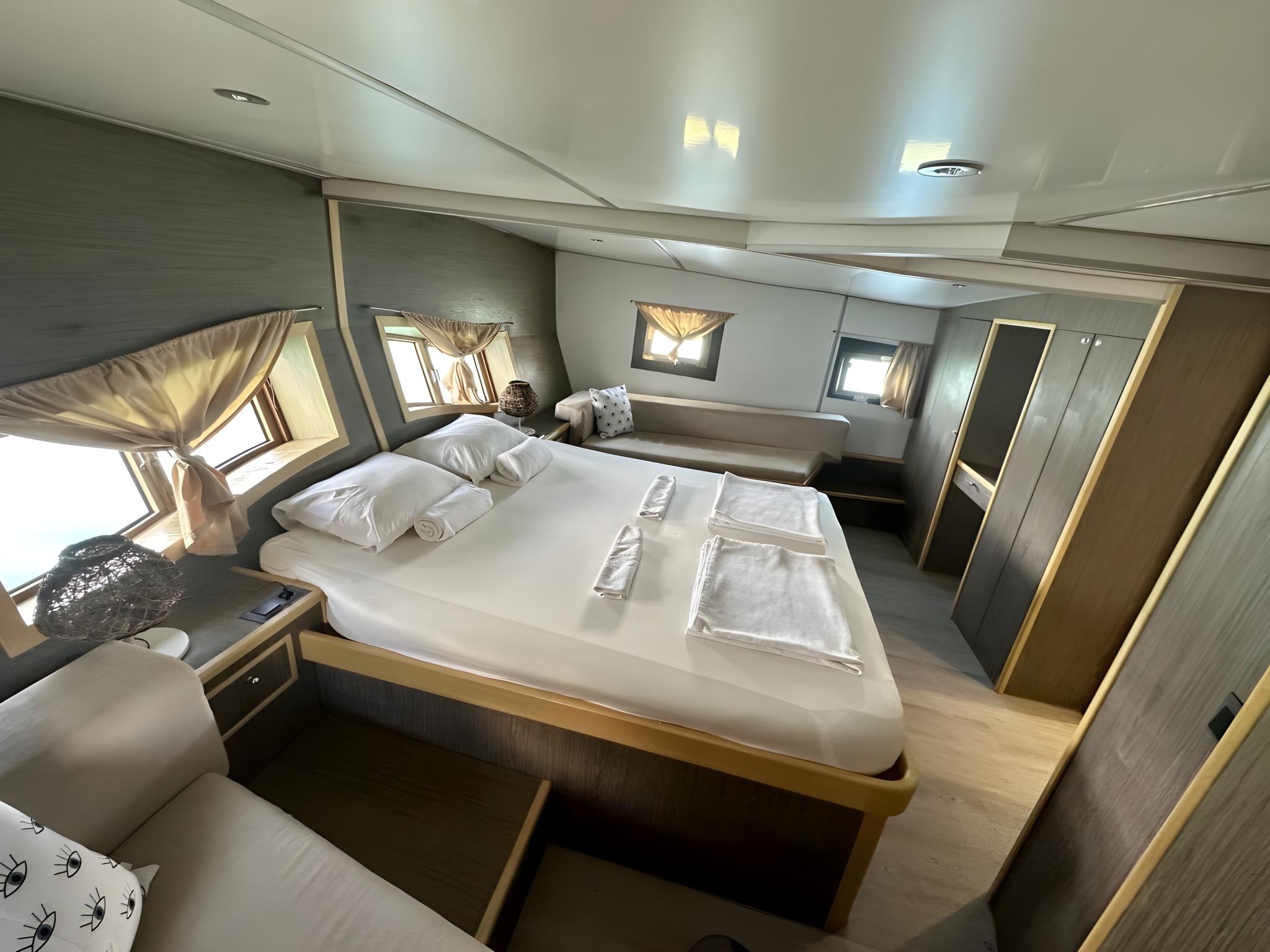 yacht cabin interior with double bed, white linens, portholes with curtains, and built-in wardrobes