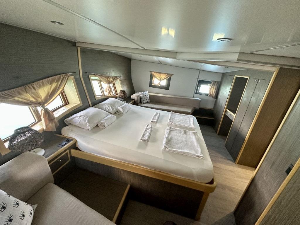 yacht cabin interior with double bed, white linens, portholes with curtains, and built-in wardrobes