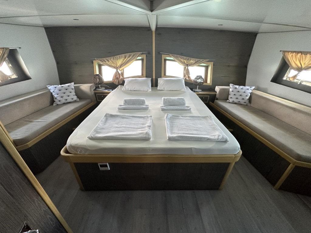yacht cabin with central double bed, built-in bench seating, portholes with curtains and folded towels