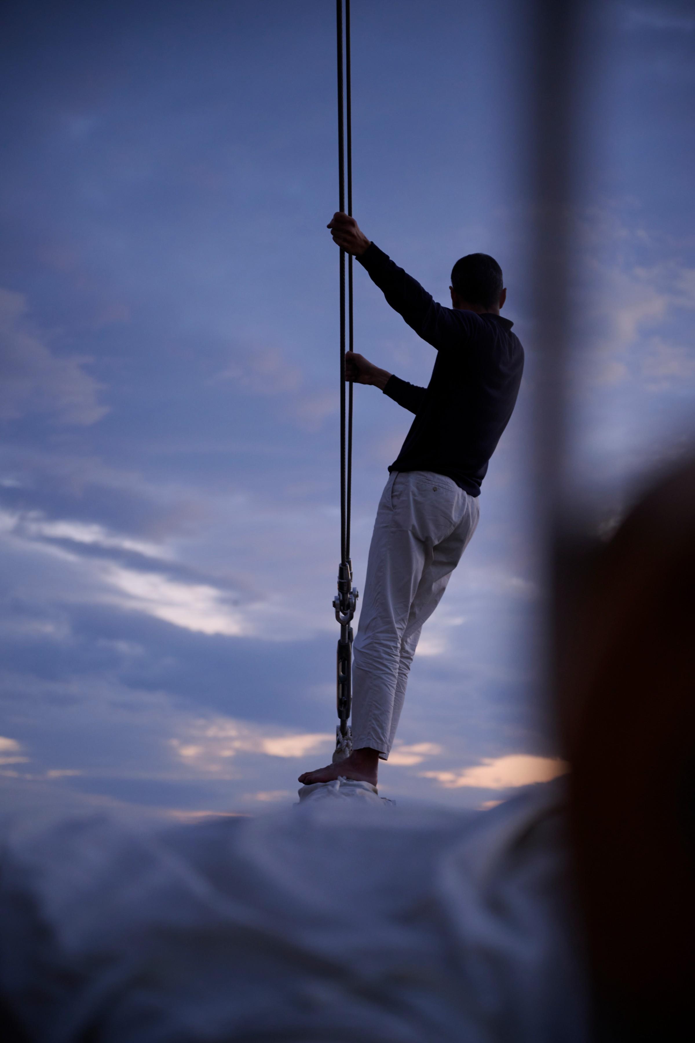 barefoot crew member gripping rigging lines on gulet bow at dusk with blue sky