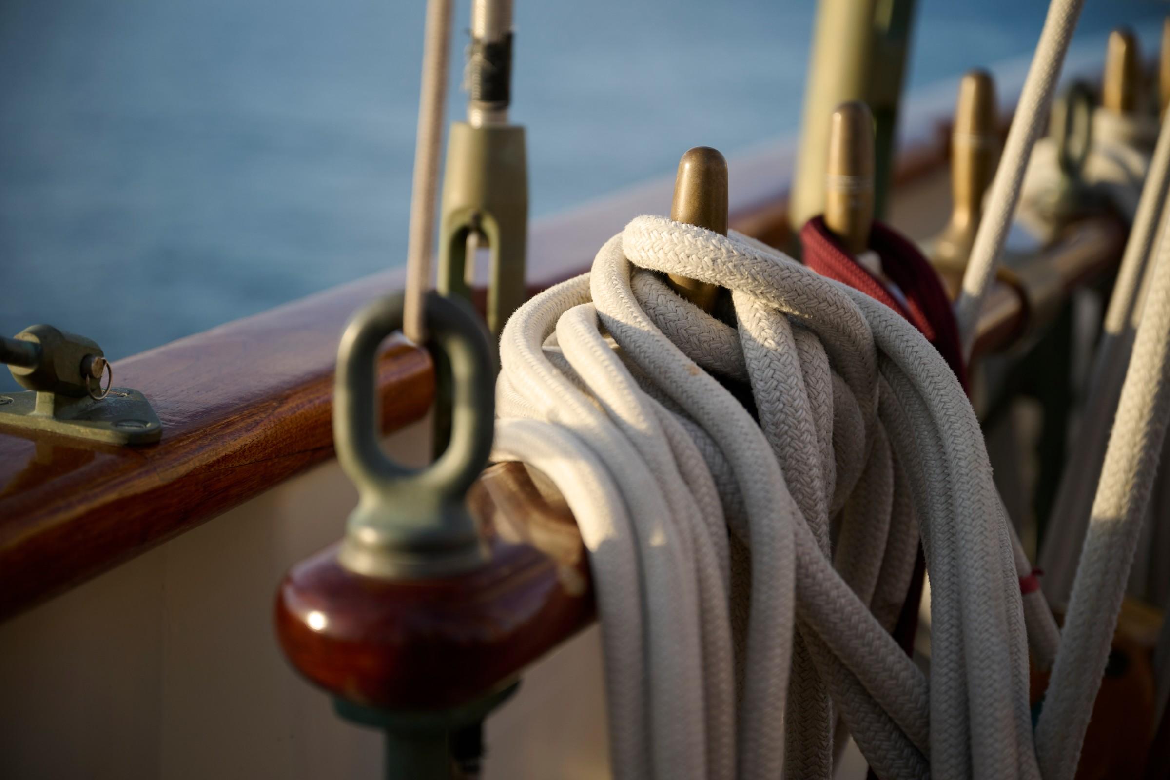 Close-up of coiled white braided ropes on polished brass belaying pins along varnished teak deck rail