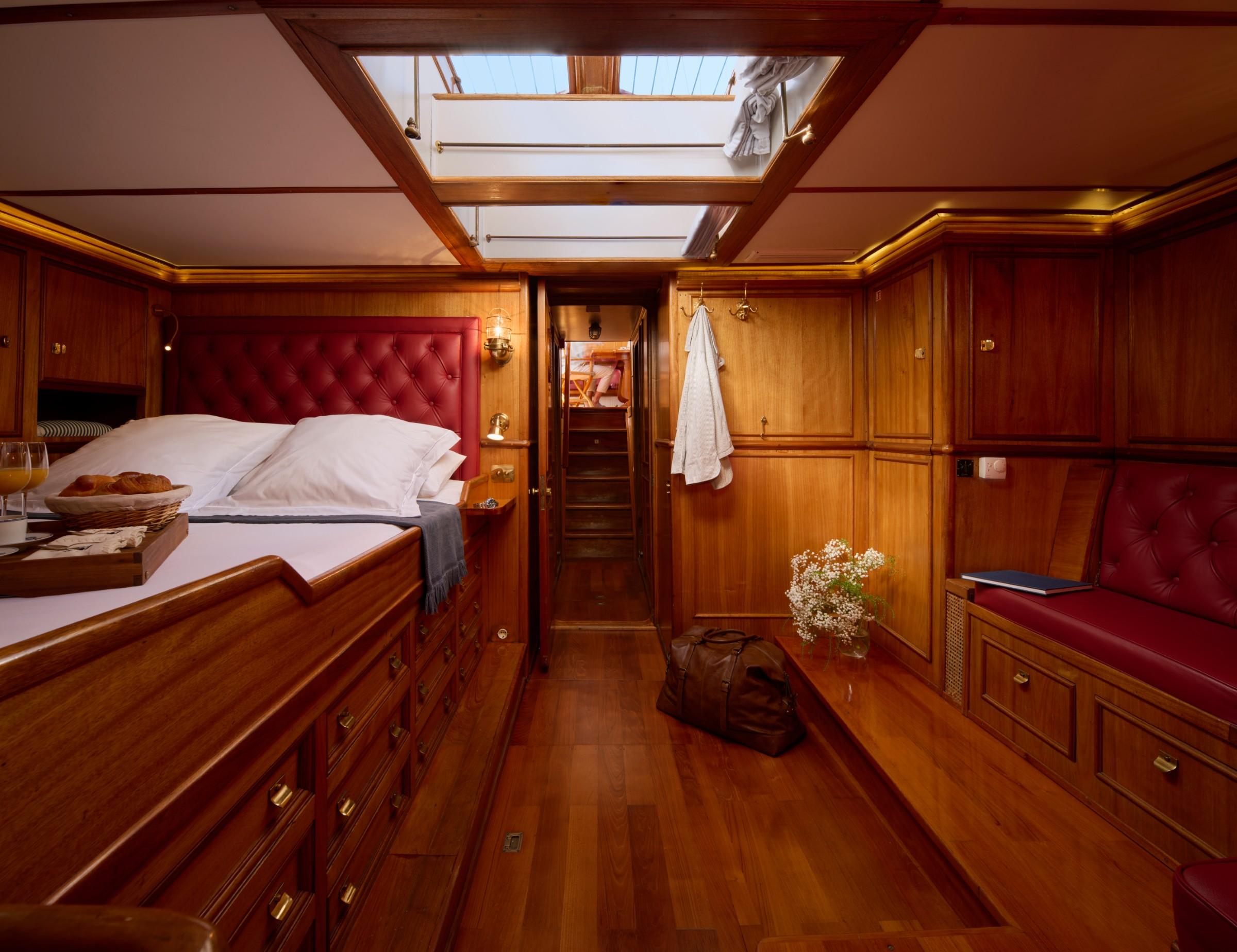 Master cabin with tufted red leather headboard, double bed, brass fittings, skylight hatch, and built-in wooden drawer units