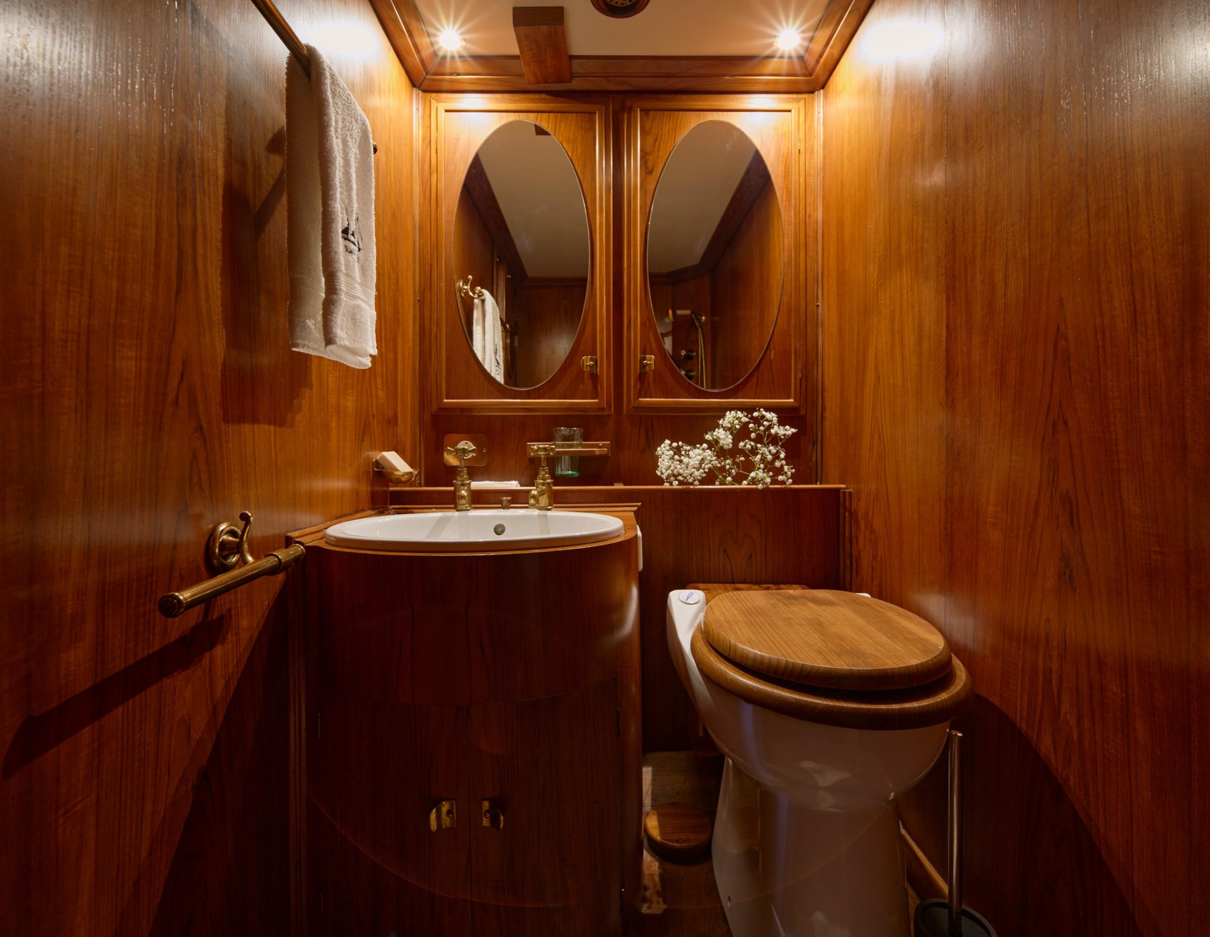 Yacht cabin bathroom with teak panelling, oval mirrors, brass fittings, pedestal sink and wooden toilet seat
