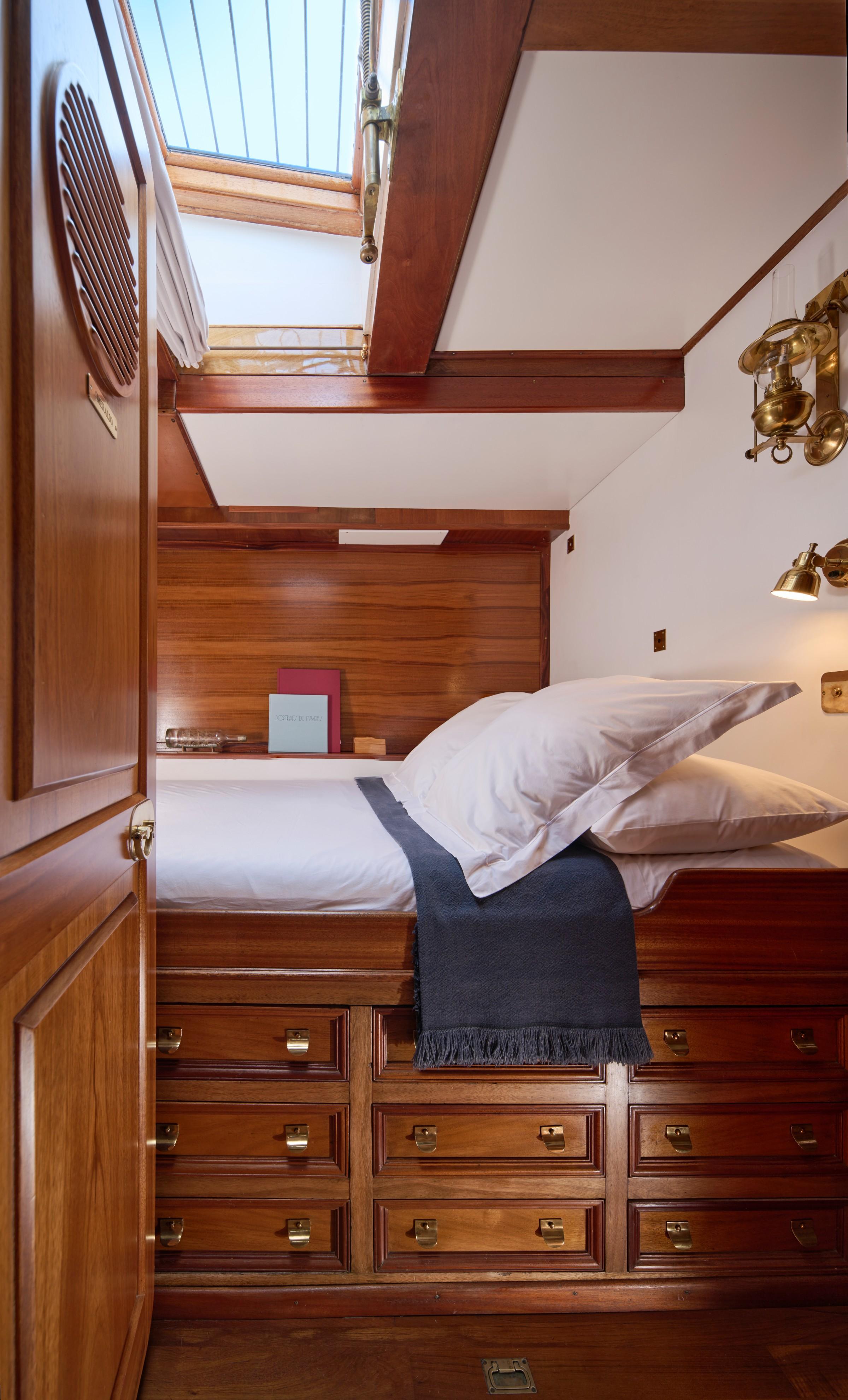 Gulet cabin with single berth, white bedding, navy throw, brass fittings, mahogany drawers and overhead skylight hatch