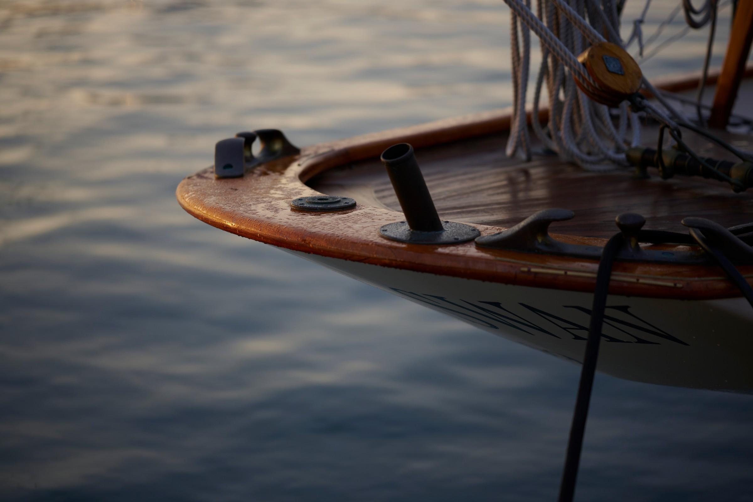 Close-up of a classic wooden sailing yacht bow with varnished teak deck, black iron chock and hawse pipe fittings, white hull, and rigging ropes at dusk