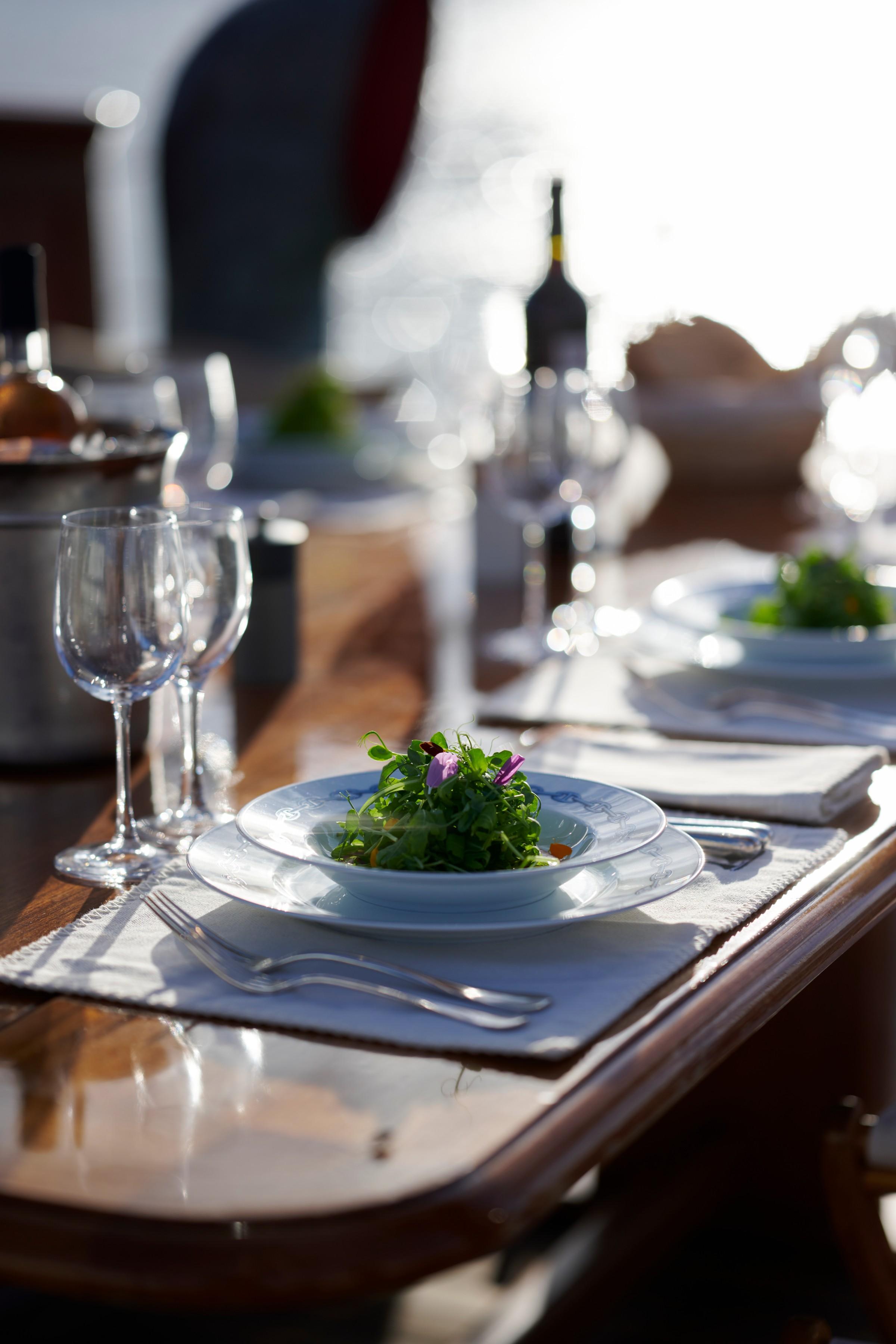 Gulet outdoor dining table set with fresh green salad garnished with edible flower, wine glasses and cutlery on white linen placemat