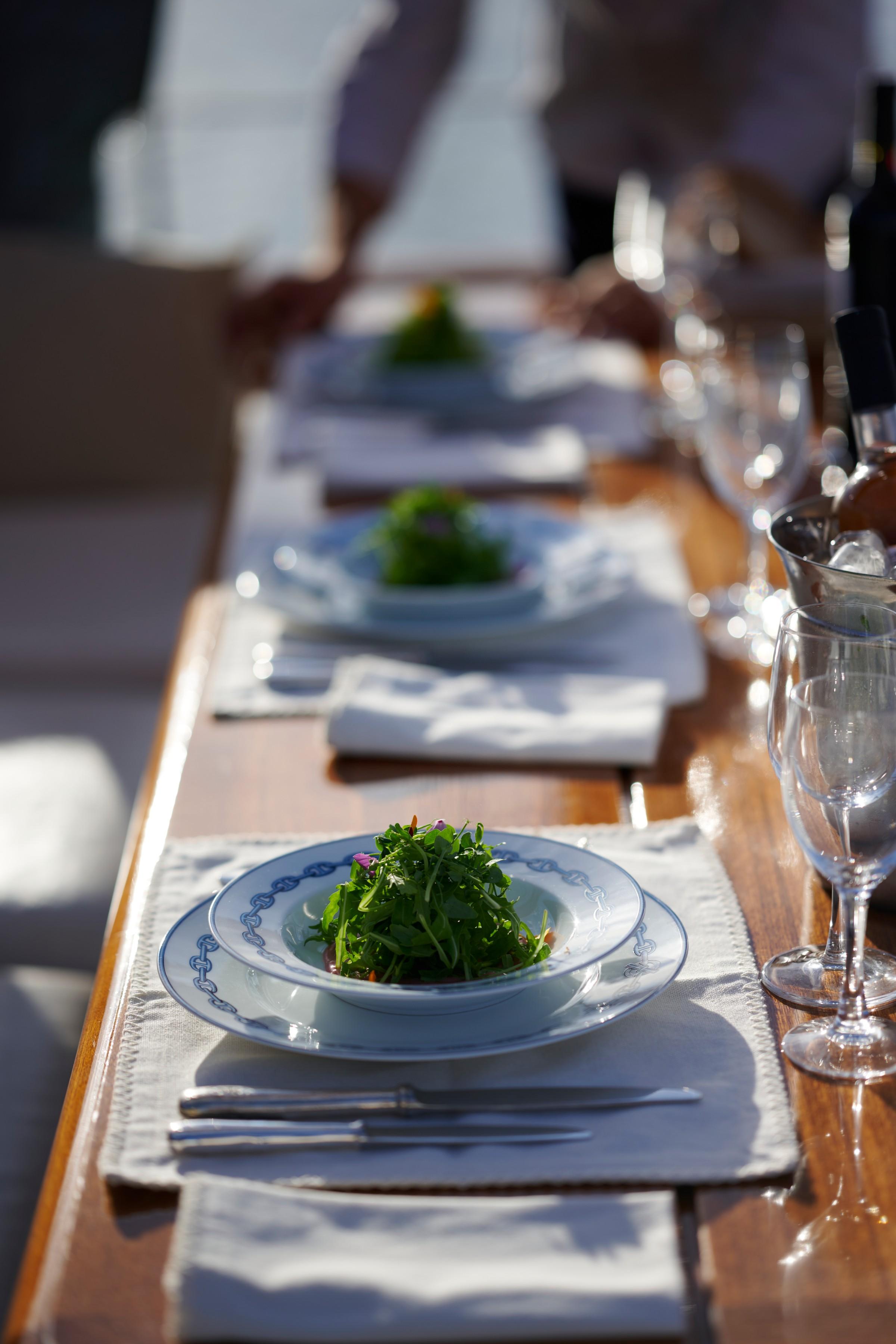 Luxury gulet outdoor dining table with green salad starters served on white and blue crested china, crystal wine glasses and teak deck