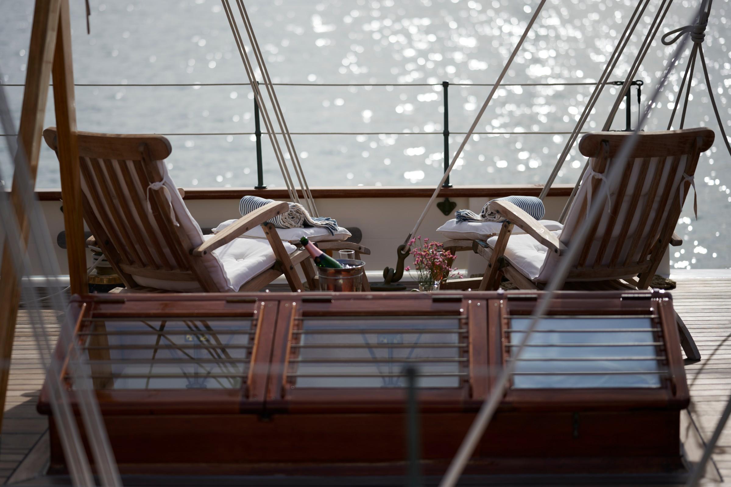 Two teak deck chairs with white cushions and champagne bucket on gulet aft deck overlooking sparkling sea