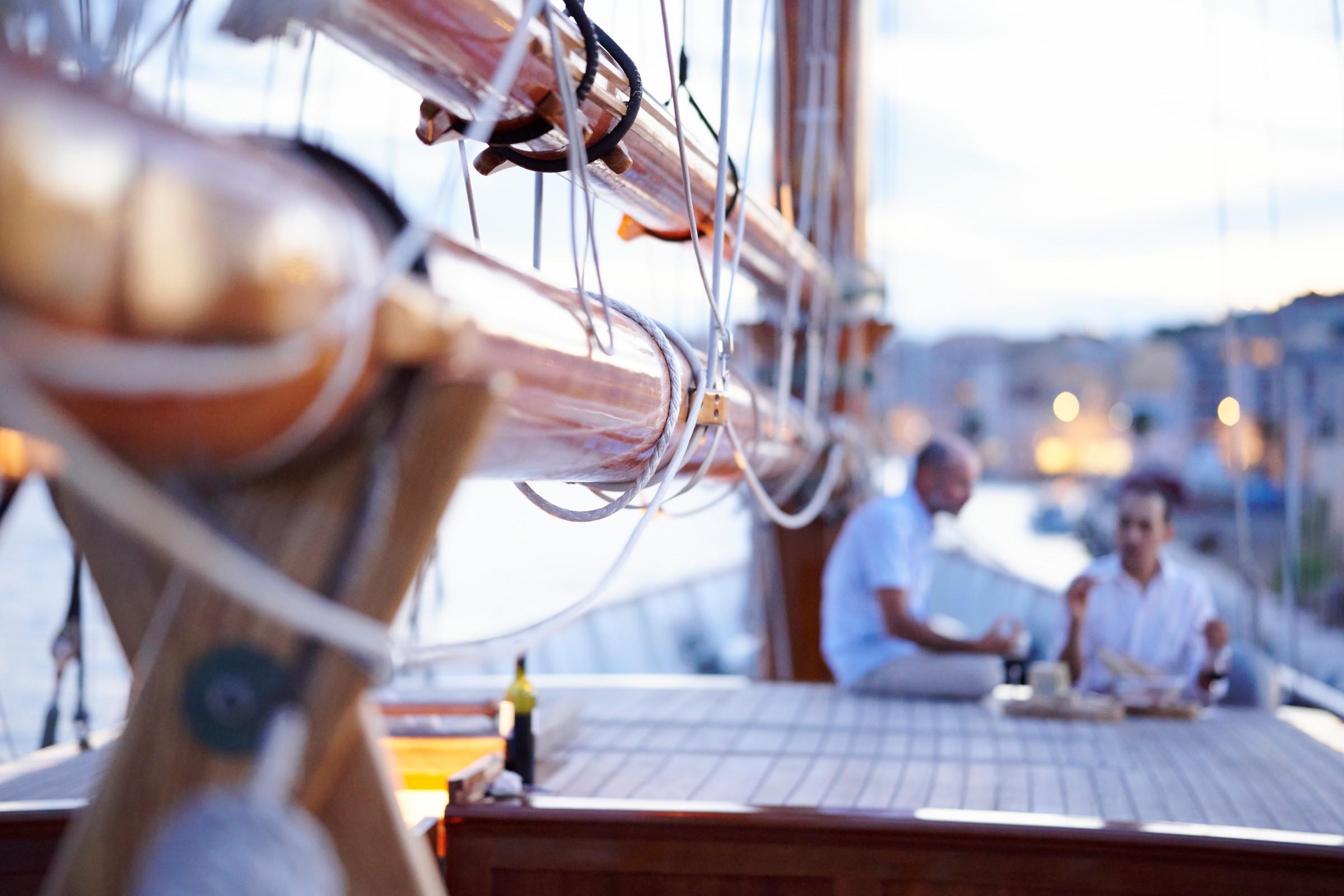 Two guests dining at teak deck table with wine bottle aboard a classic wooden gulet moored in a marina at dusk