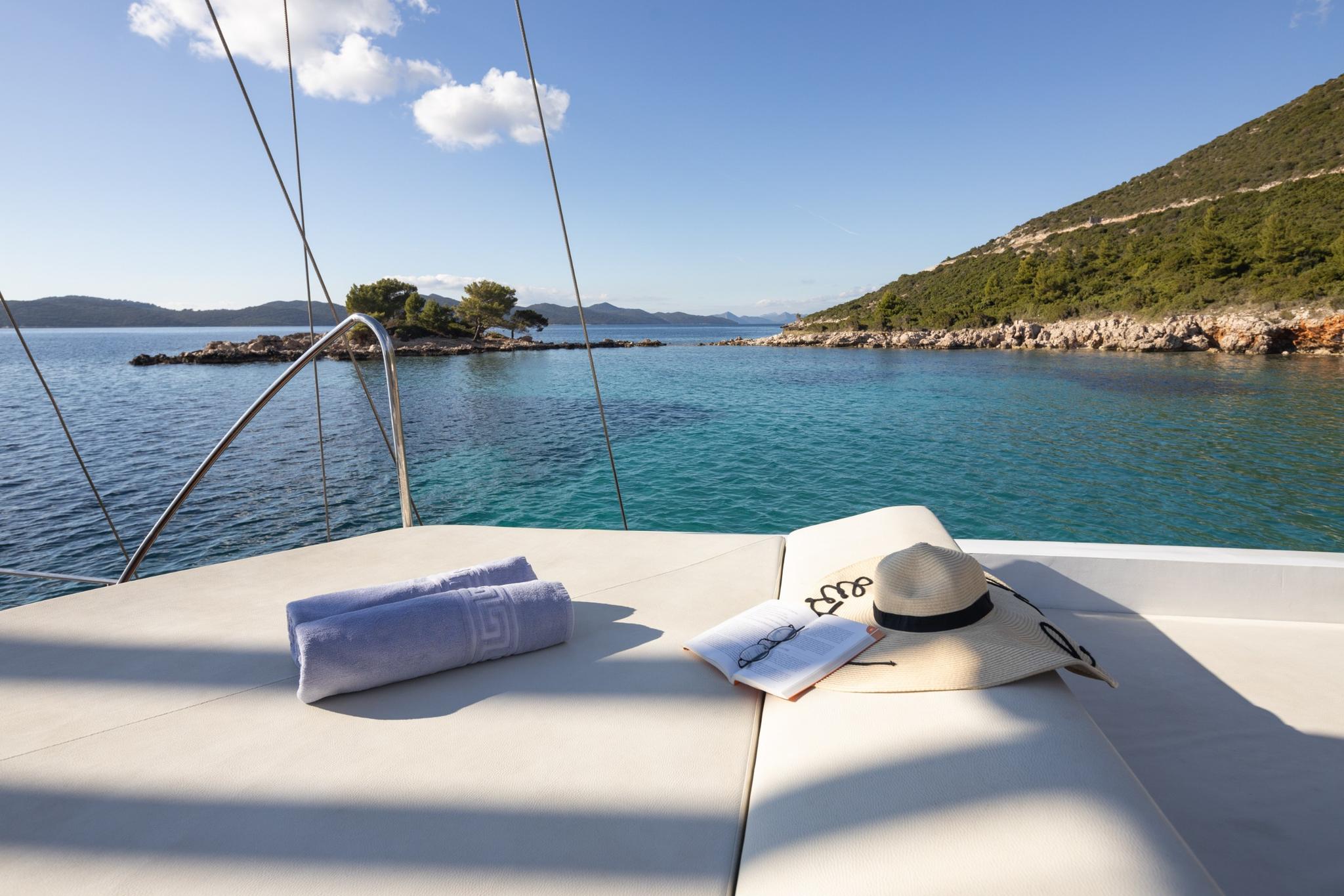 yacht bow sun deck with white cushioned seating, towels and hat, overlooking turquoise Mediterranean bay