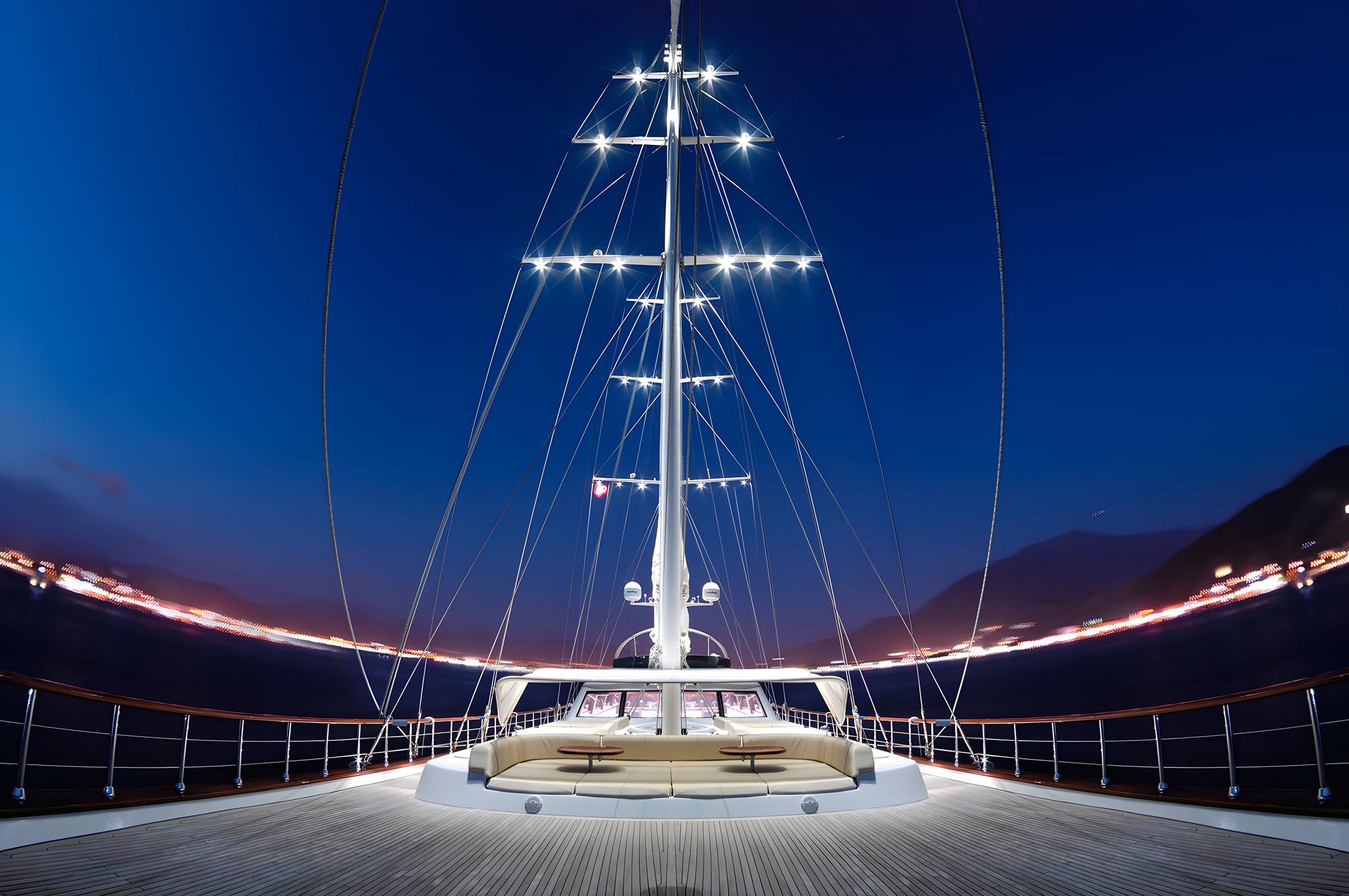 Luxury sailing yacht bow deck with curved seating area and illuminated mast at twilight