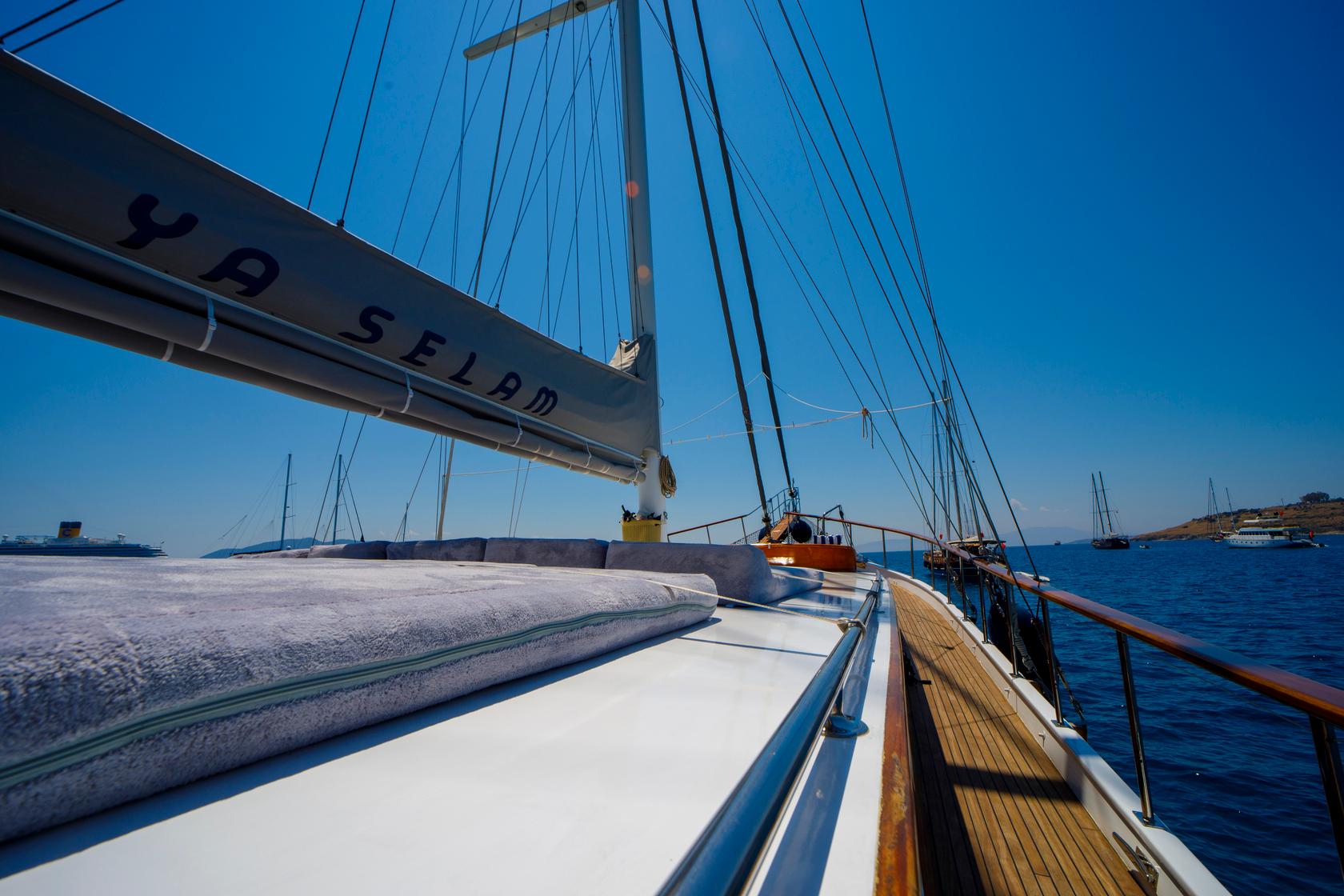 bow deck of sailing gulet showing white cushioned seating, rigging, and Mediterranean coastline