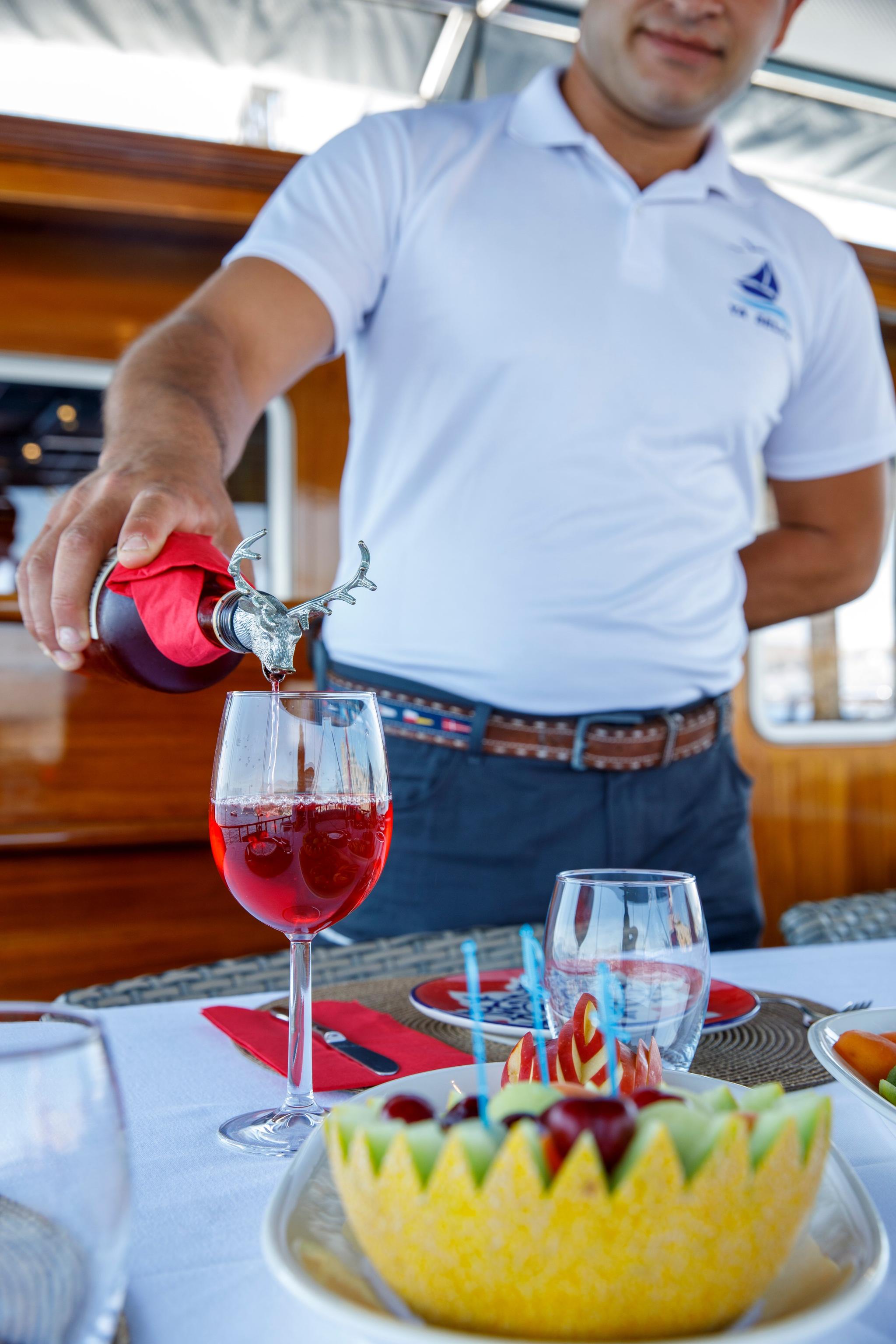 yacht crew member in white polo shirt pouring red wine into glass with fruit bowl on dining table