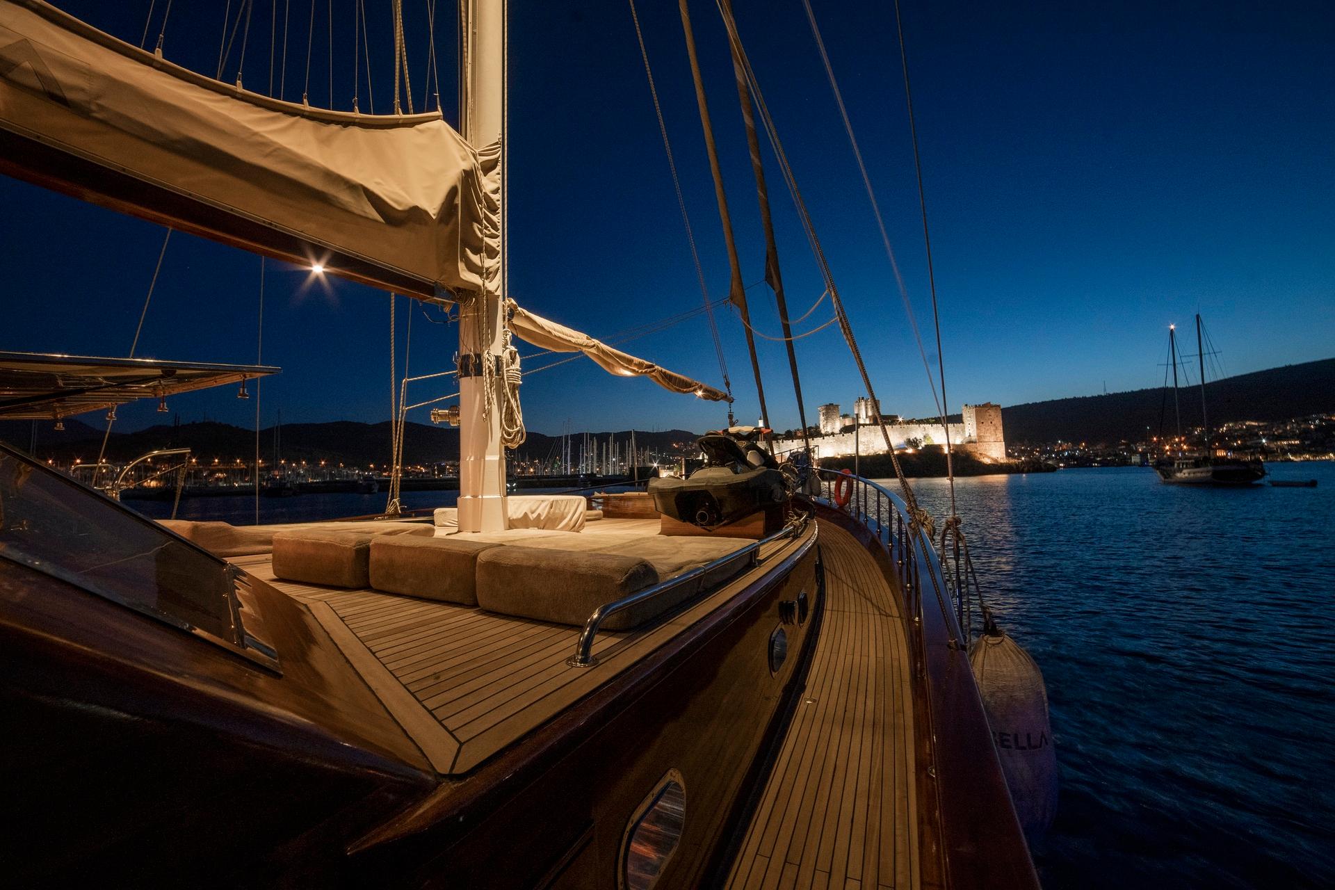 gulet deck with beige cushioned seating area overlooking illuminated harbor castle at twilight