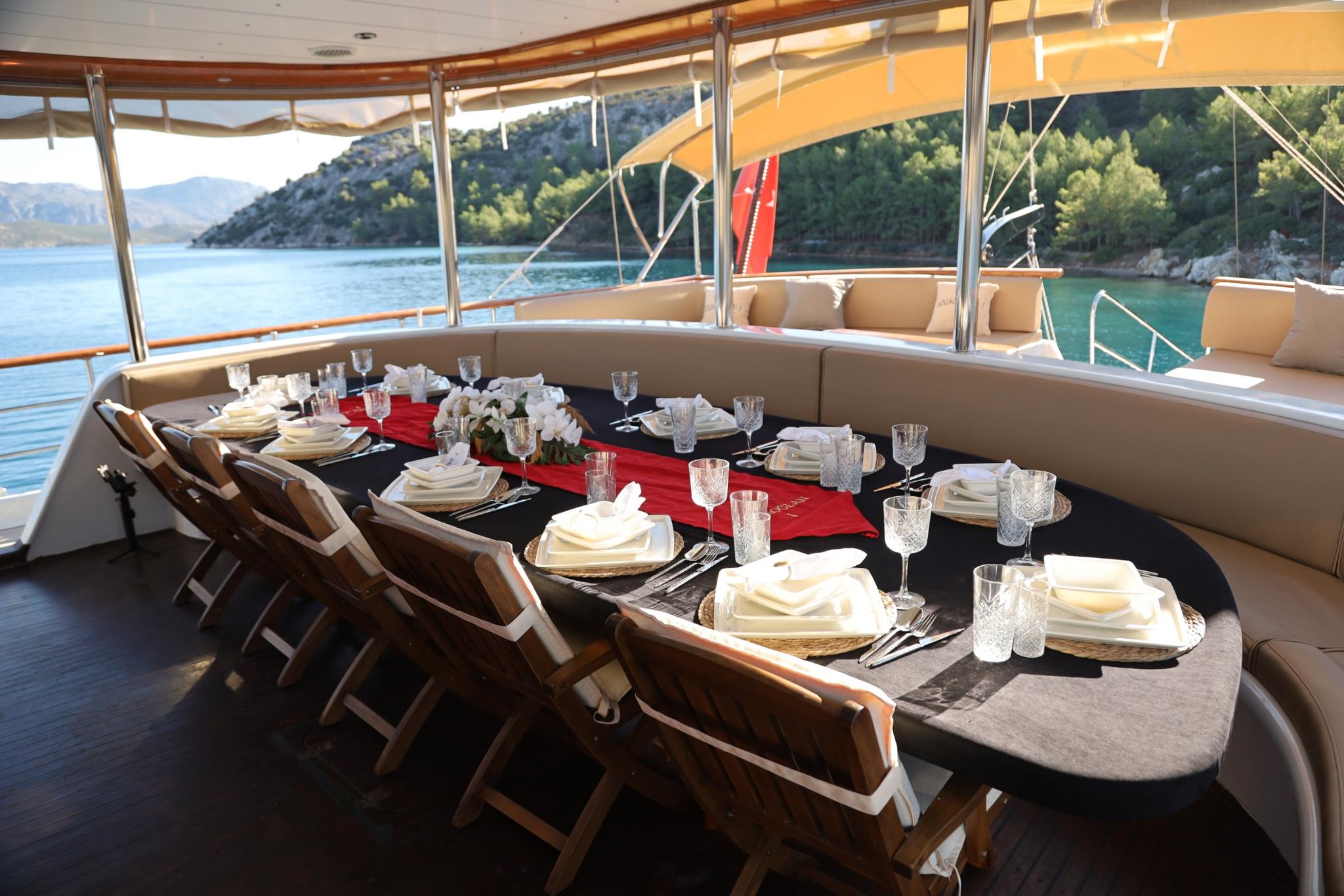 Aft deck dining area with oval table set for formal dinner, curved seating and mountain coastline views