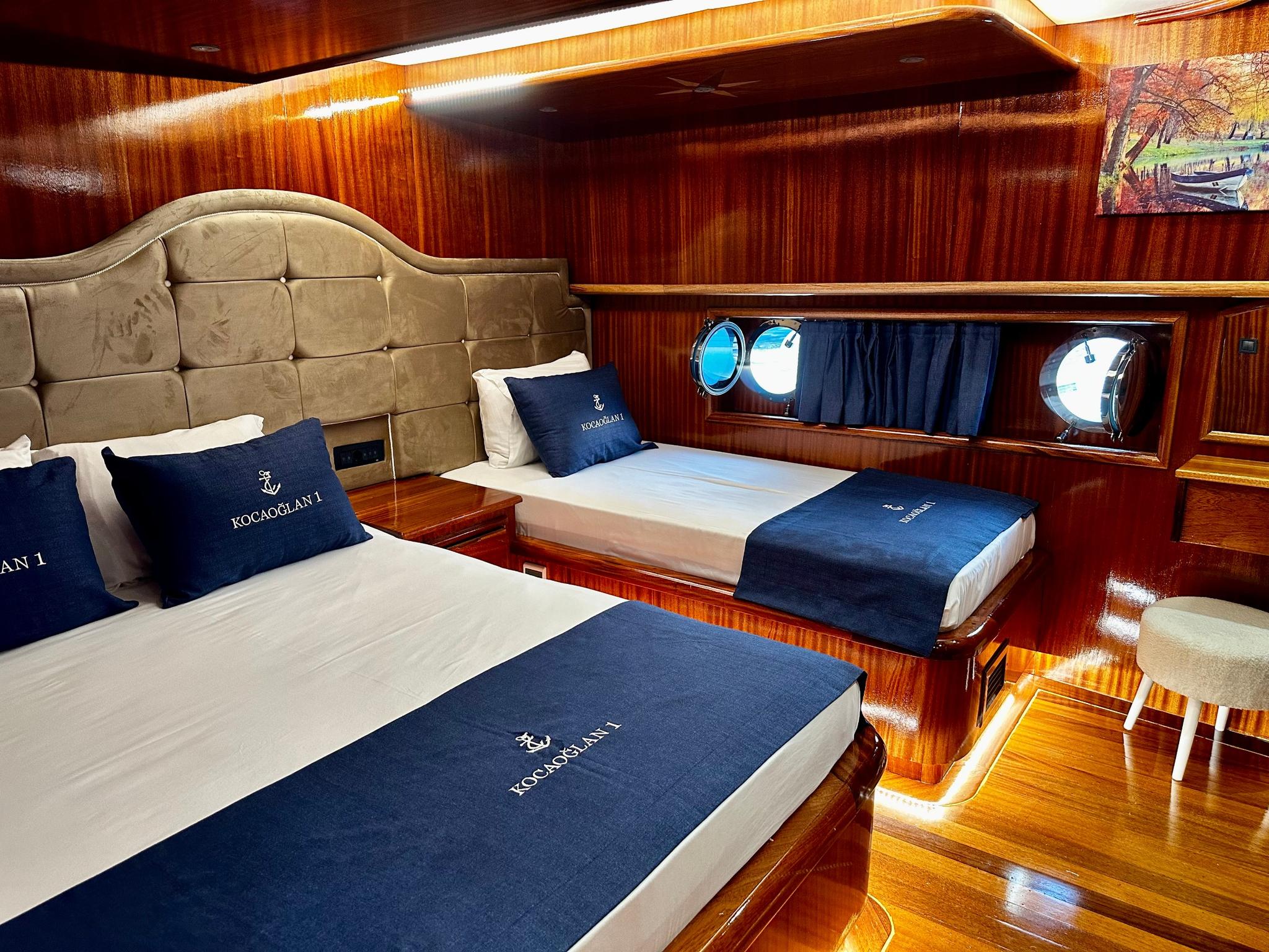 yacht twin cabin with beige upholstered headboard, navy KOCAOGLAN branded bedding and portholes